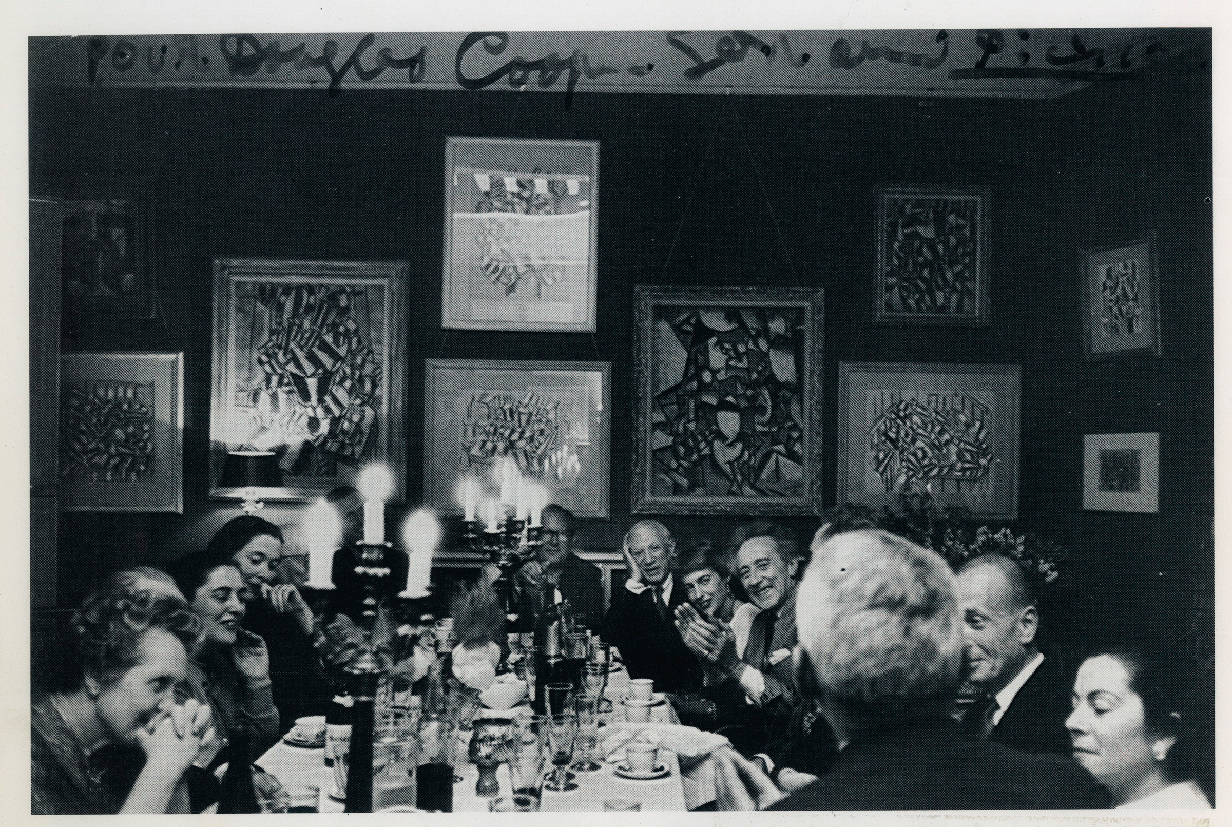 The image shows a vintage black and white photo of a group of people at a dinner party, surrounded by walls adorned with various framed artworks.