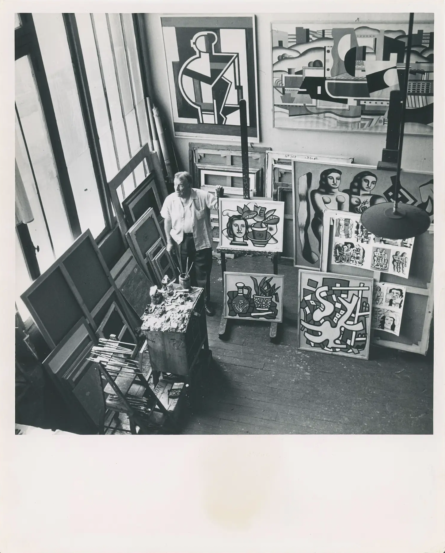 An artist stands in a studio filled with abstract and cubist-style paintings and art supplies.