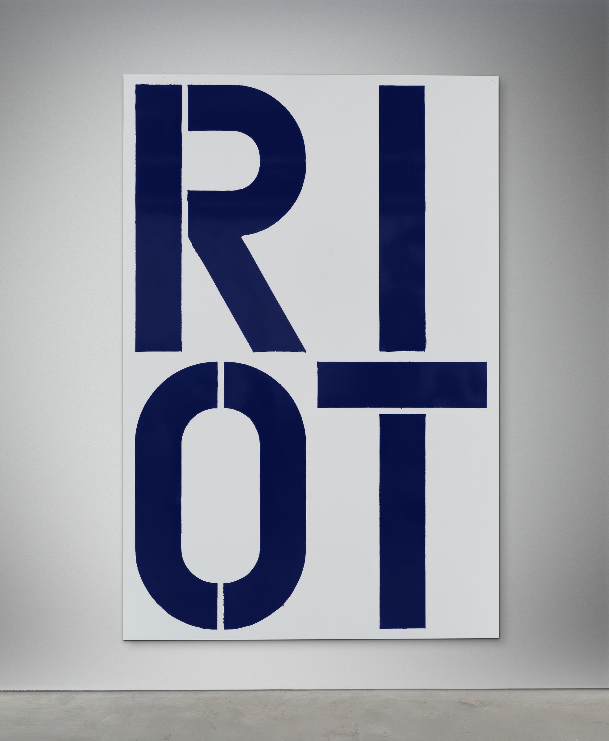 A painting titled "RIOT" using bold blue letters on a white canvas, displayed against a gray wall.