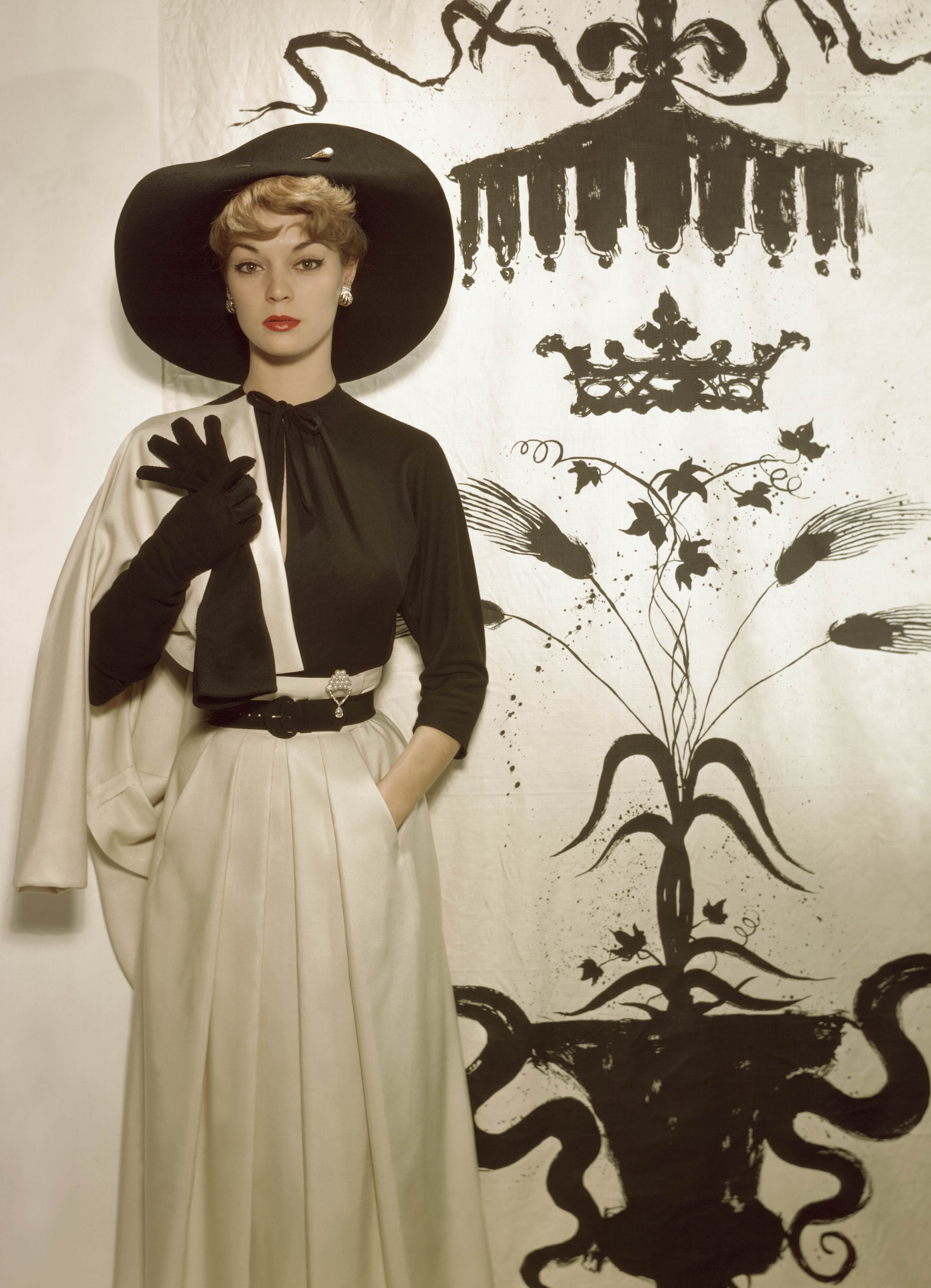 A person in elegant vintage attire poses against a backdrop featuring intricate black crown and floral designs.