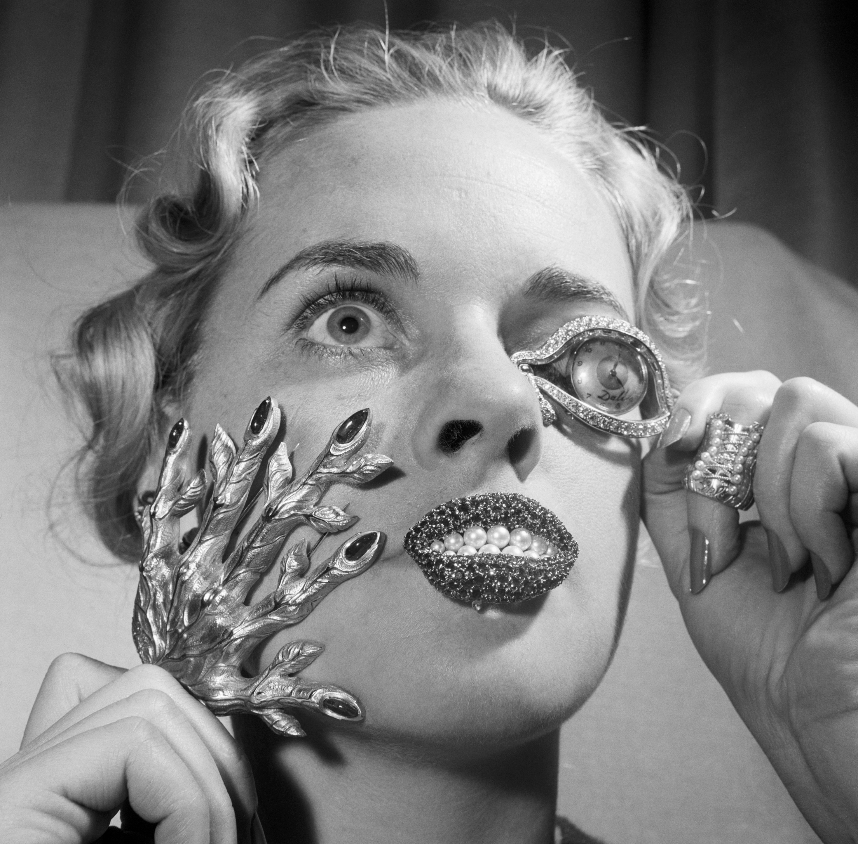 A person with light wavy hair holds a silver textured object to each ear and wears a chunky metallic ring on one finger.