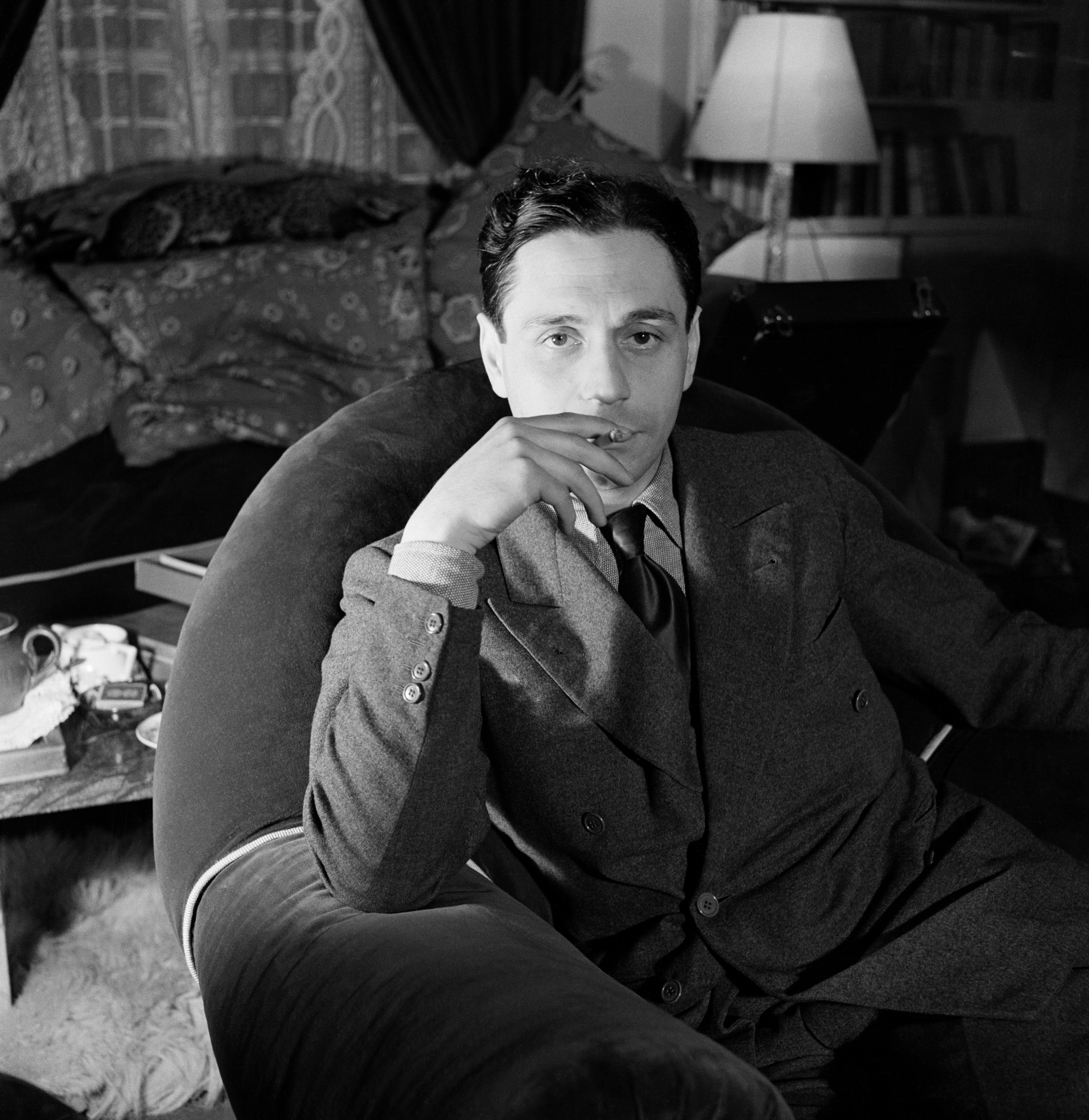 A person in a suit sits on a curved chair in a cozy, dimly lit living room with a lamp, books, and patterned cushions.