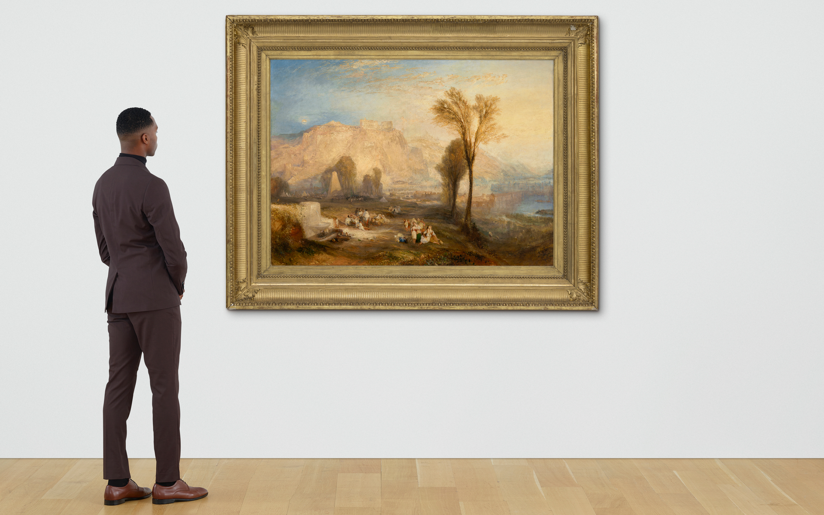 A person in a dark suit views a large framed landscape painting in a gallery with light wooden flooring.