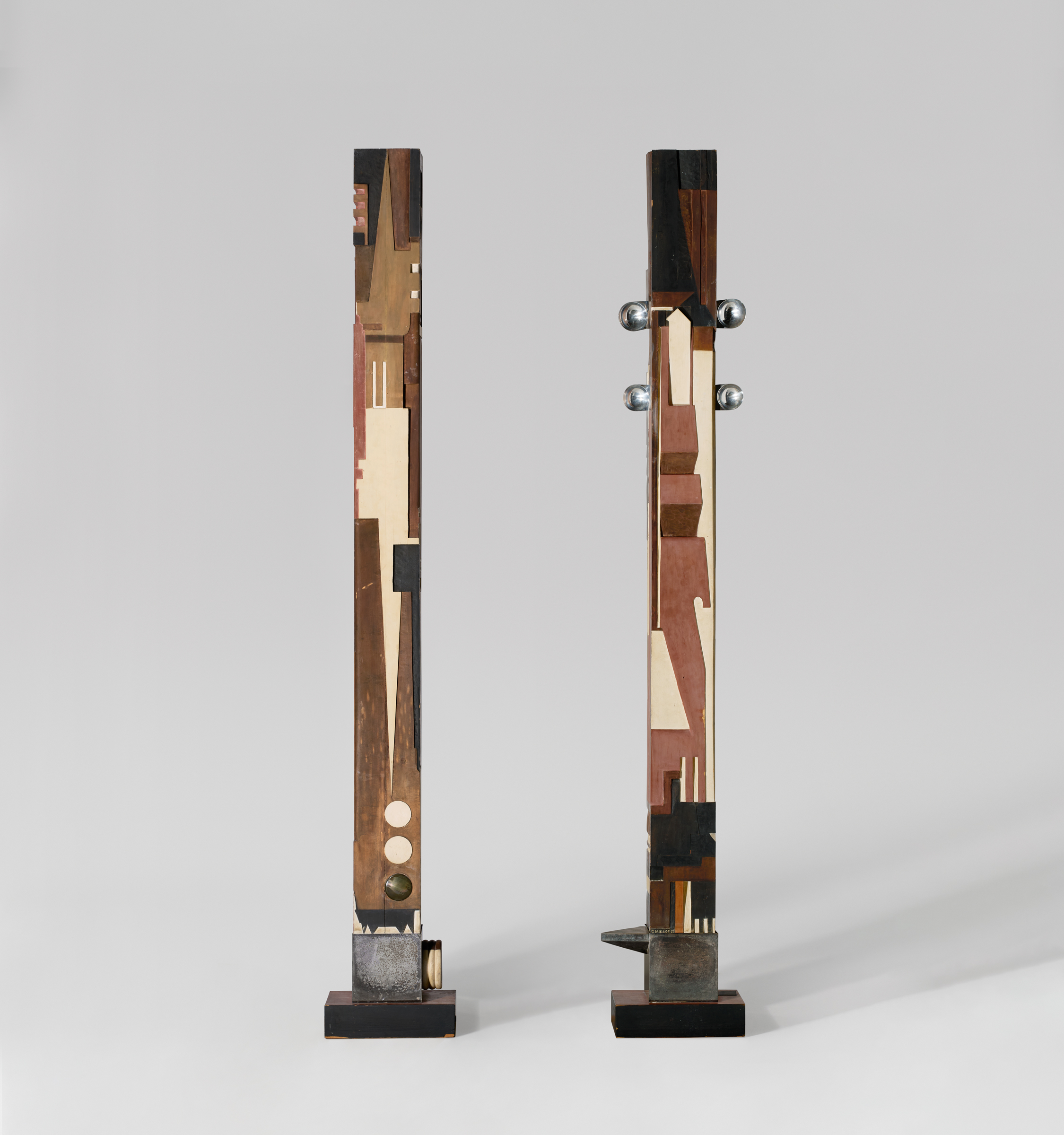 Two tall, narrow wooden sculptures with geometric patterns and metal accents stand on black bases against a plain gray background.