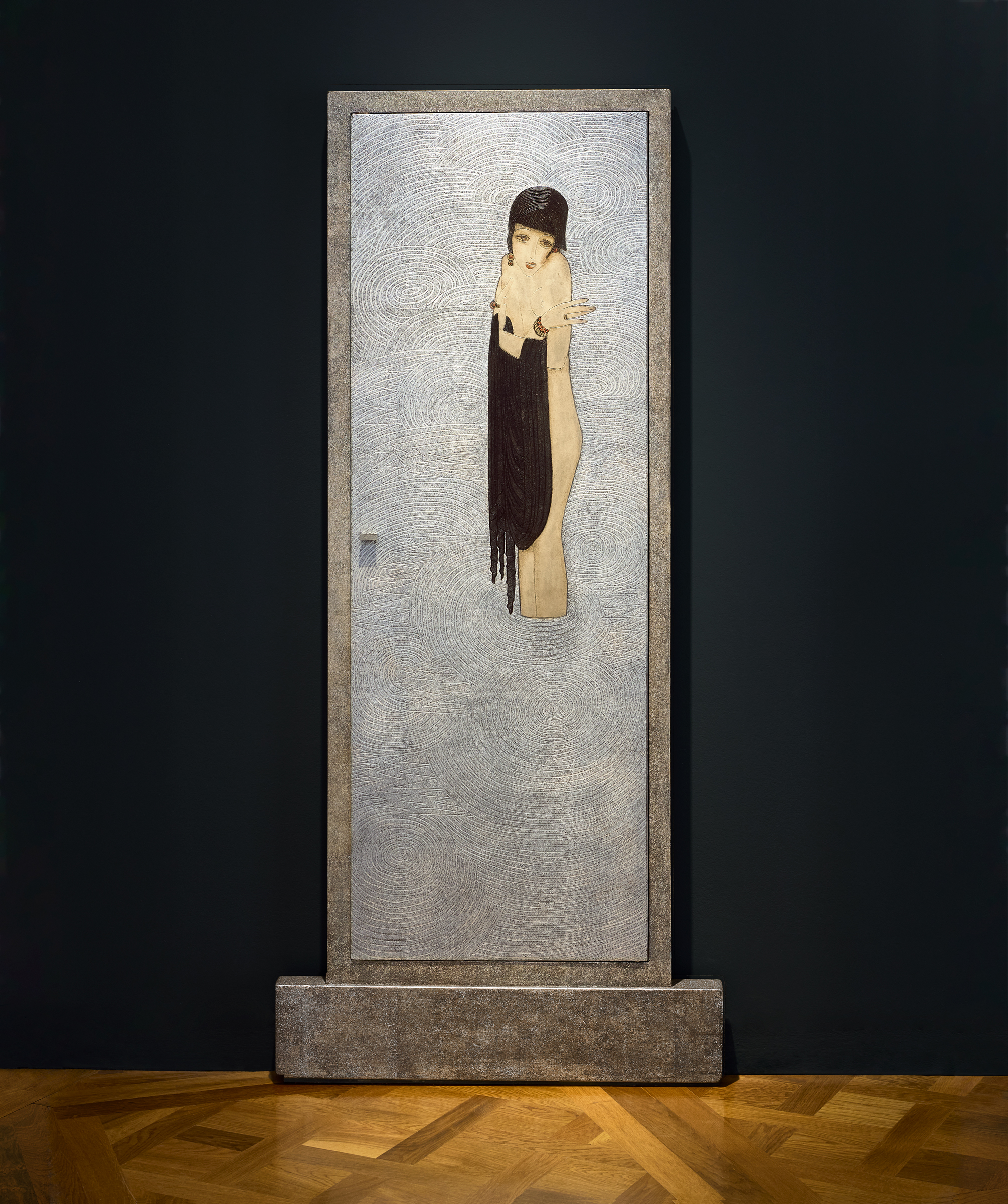 A mixed-media sculpture of a figure with long dark hair stands against a textured rectangular background on a wooden floor.