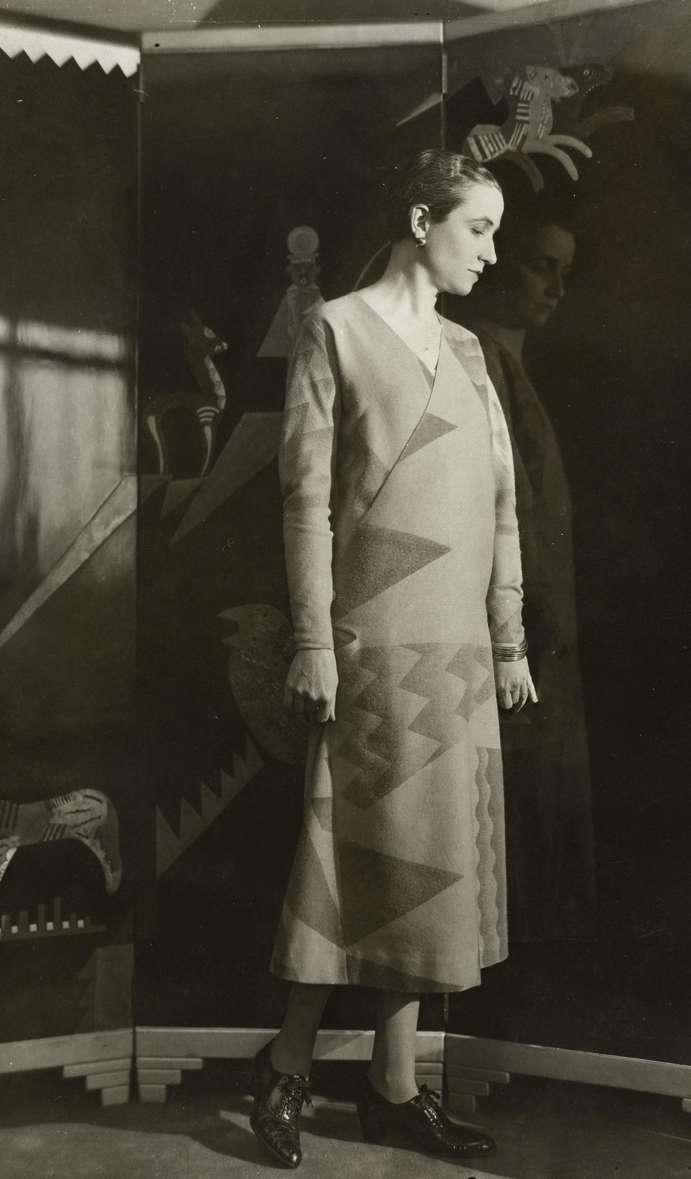 A person stands in front of a painted backdrop, wearing a geometric-patterned dress and dark shoes.