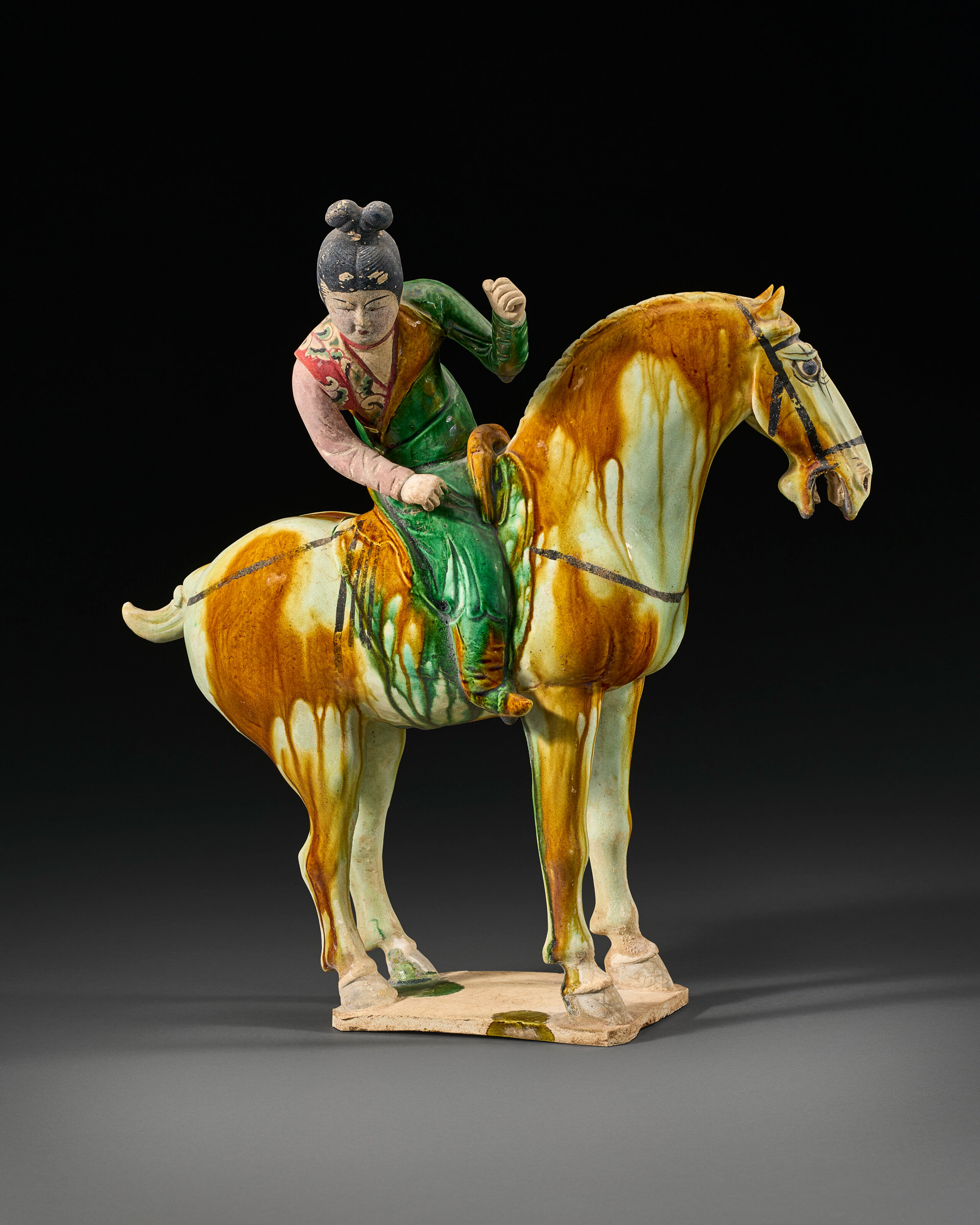 A glazed ceramic figure of a rider wearing green and pink, mounted on a yellowish-brown horse.