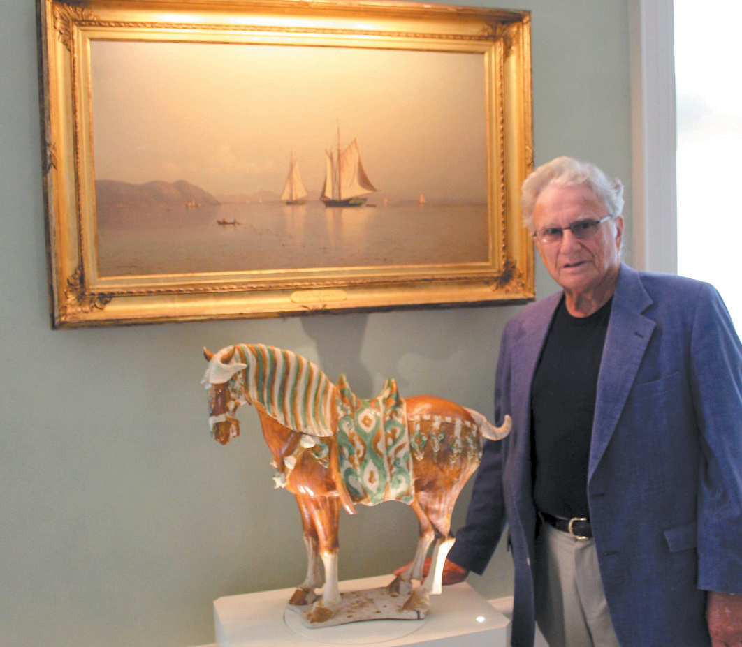 A person in a blue blazer stands beside a ceramic horse statue beneath a framed painting of ships.
