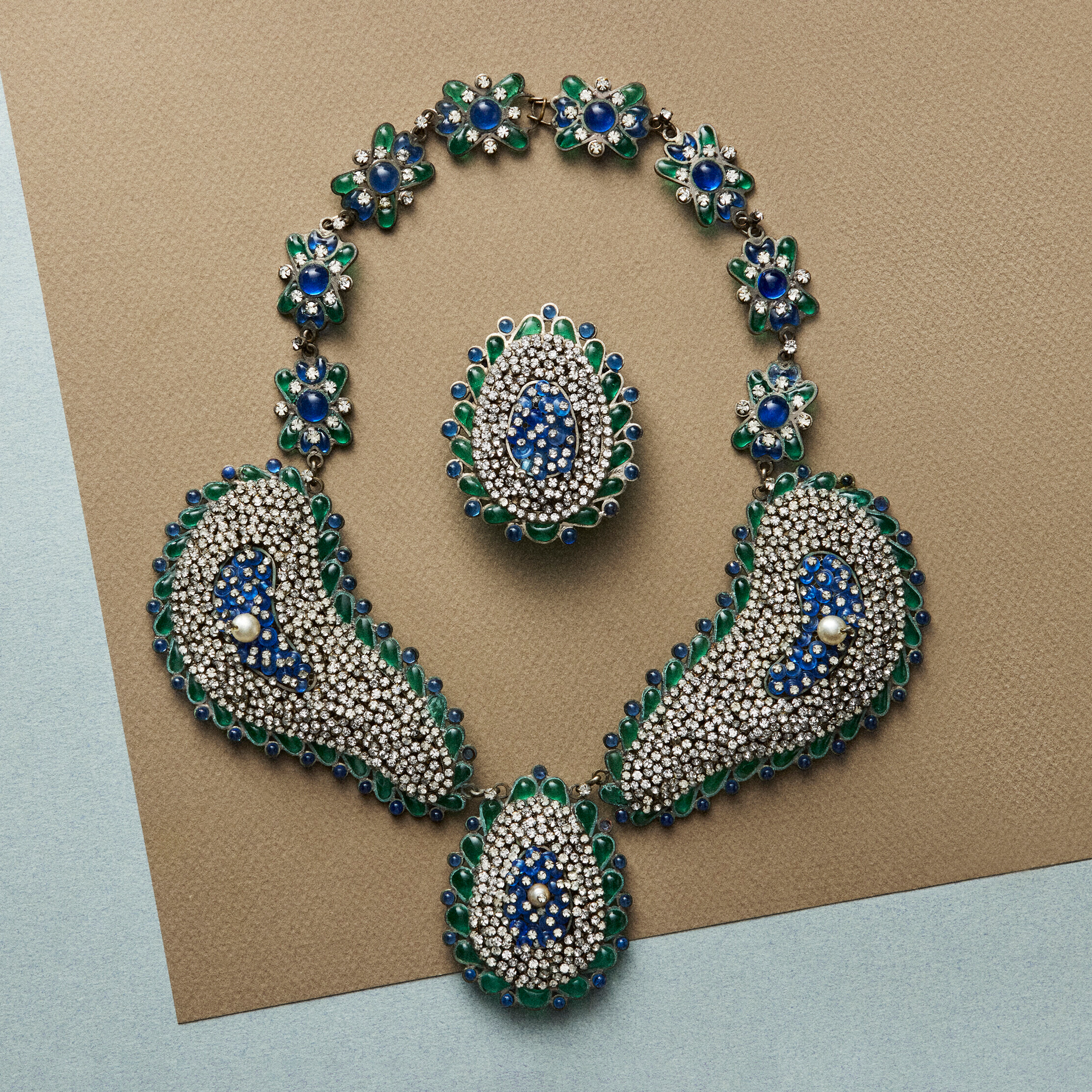 A jeweled necklace and ring set featuring blue, green, and silver stones arranged in paisley and floral designs on a neutral background.