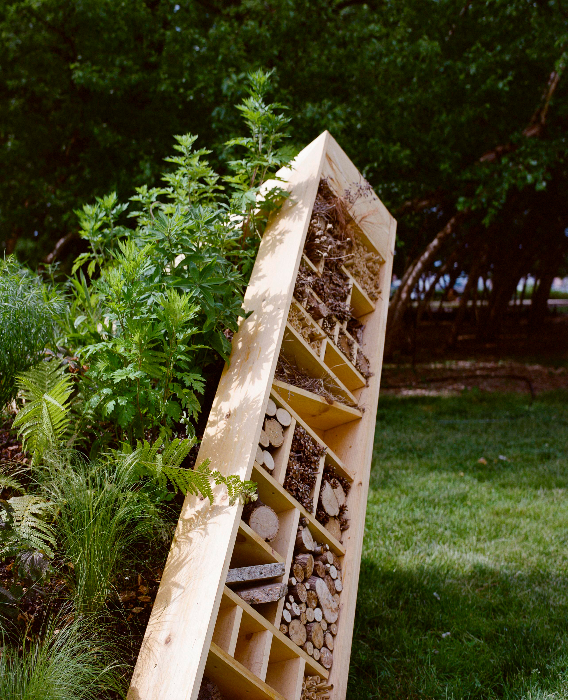 A wooden insect hotel filled with logs, sticks, and plants stands on grass near leafy greenery.