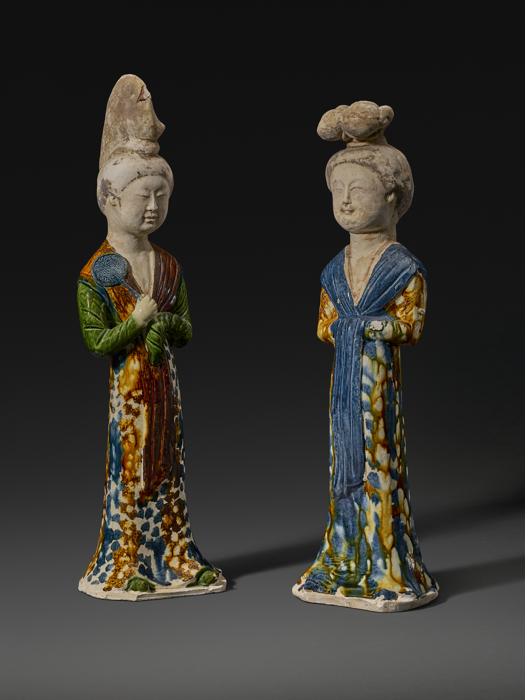 Two colorful, elongated ceramic figurines in patterned robes stand against a dark background.