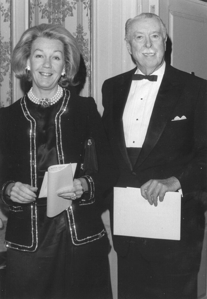 A woman in formal attire with pearls stands beside a man in a tuxedo holding papers.