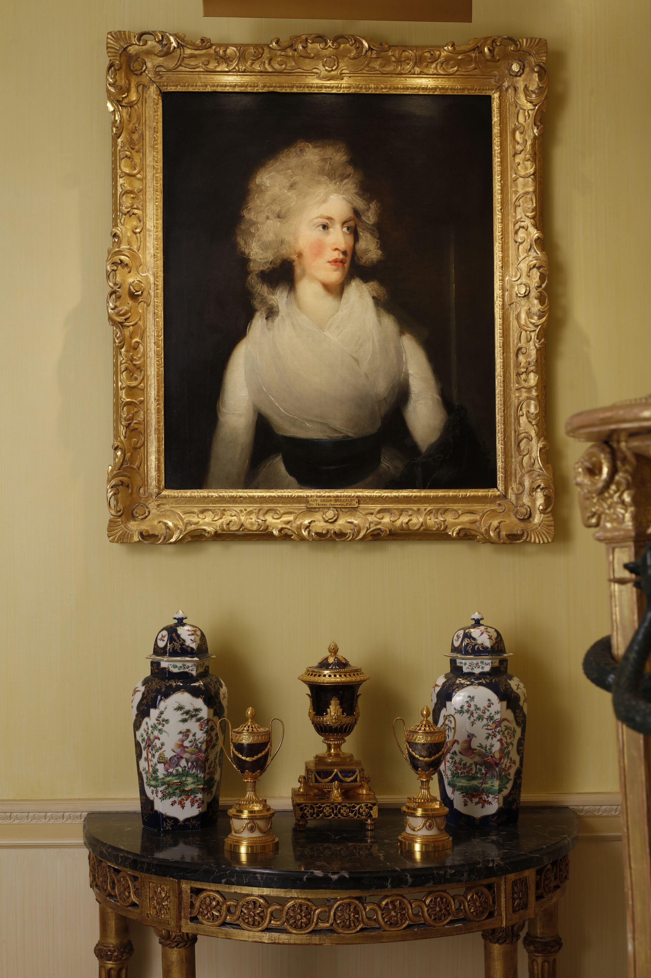 A classic interior features a gilded portrait above a marble-topped console with ornate vases and urns.