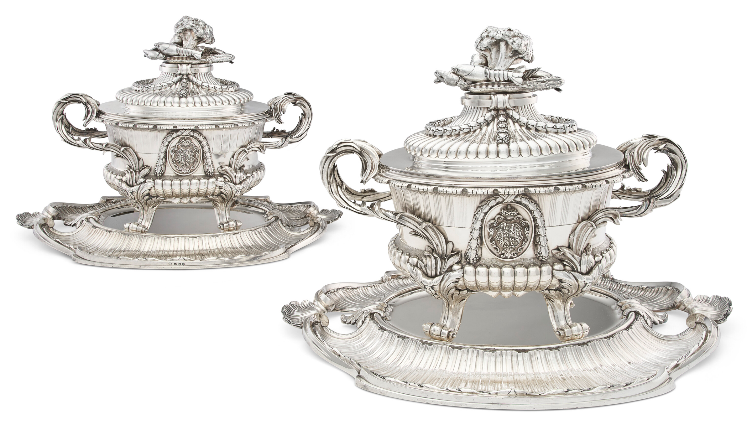 Two ornate, silver soup tureens with matching trays and decorative handles and lids.