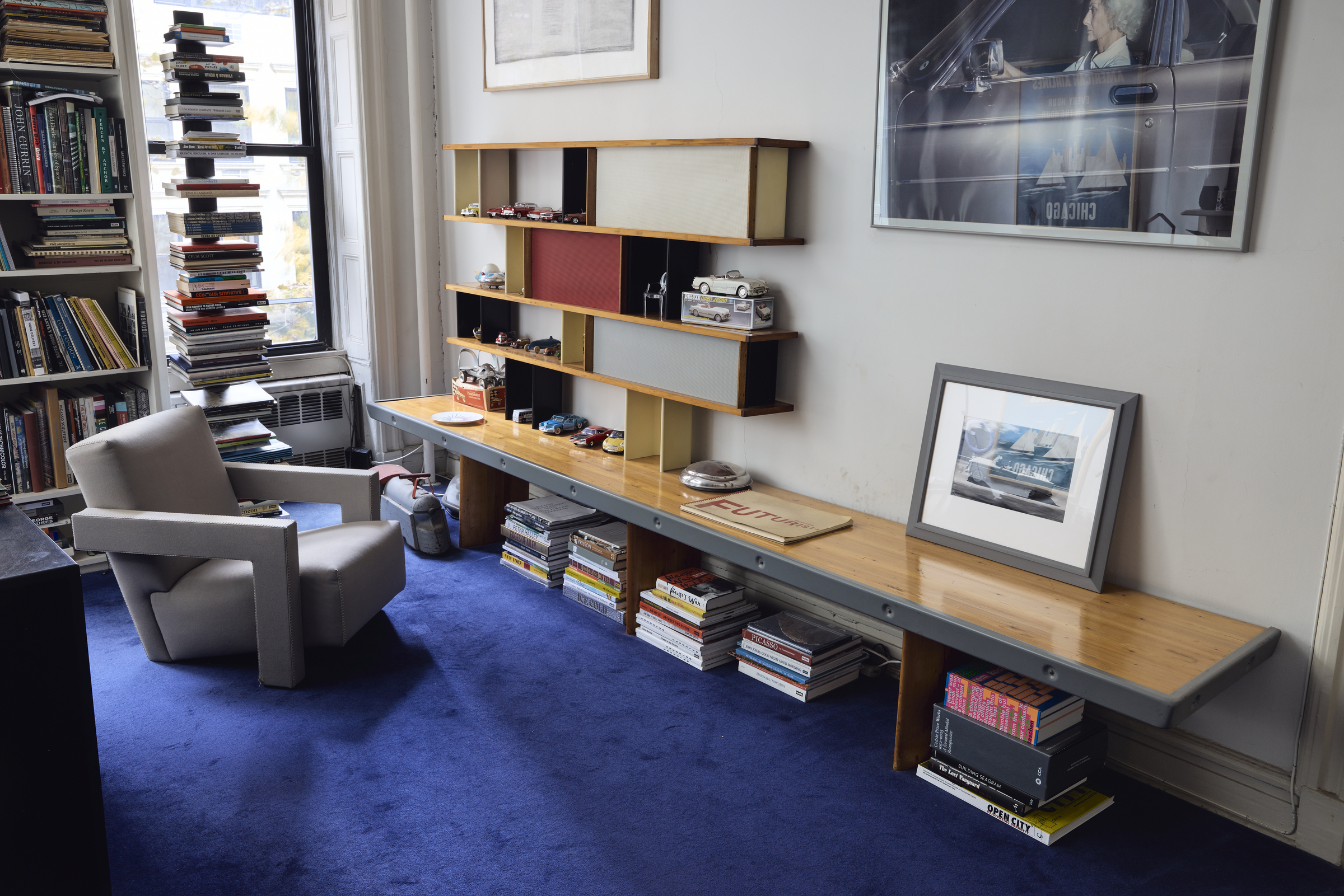 A cozy room features bookshelves, a modern chair, a blue carpet, and model cars on display.