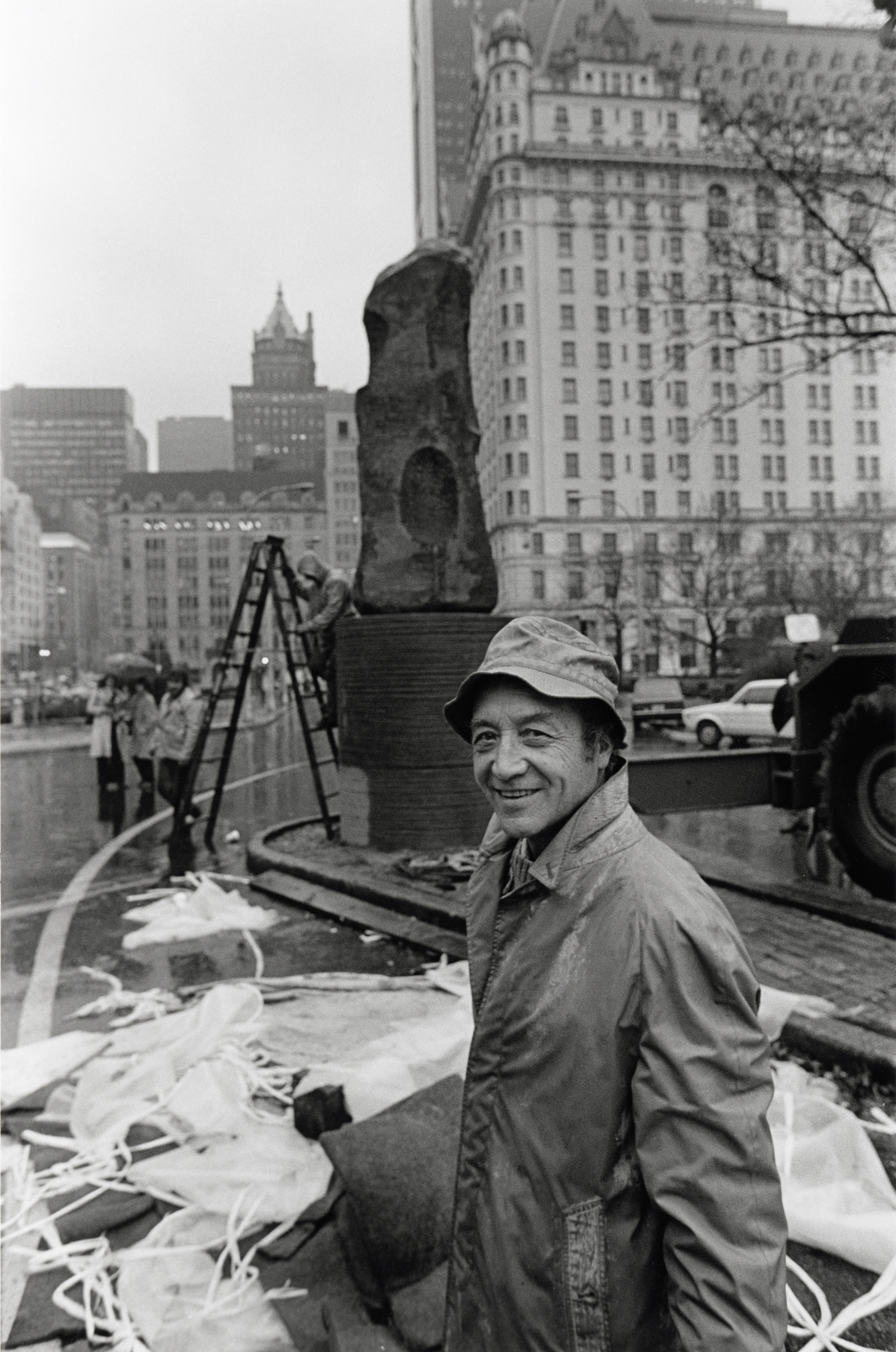 A person in a raincoat stands near an abstract sculpture and construction materials in a city.