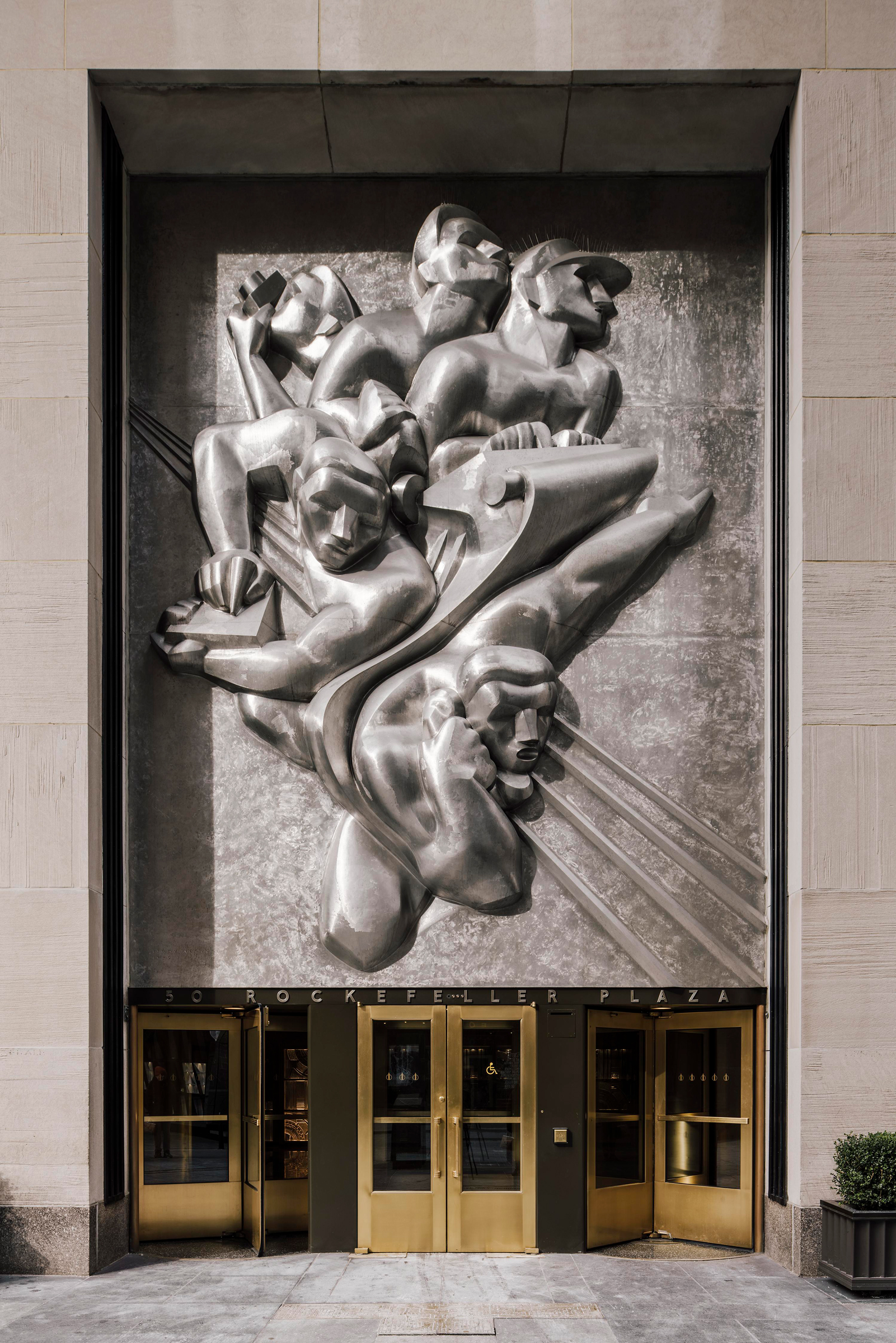 A metallic Art Deco relief sculpture is displayed above golden entrance doors at Rockefeller Plaza.