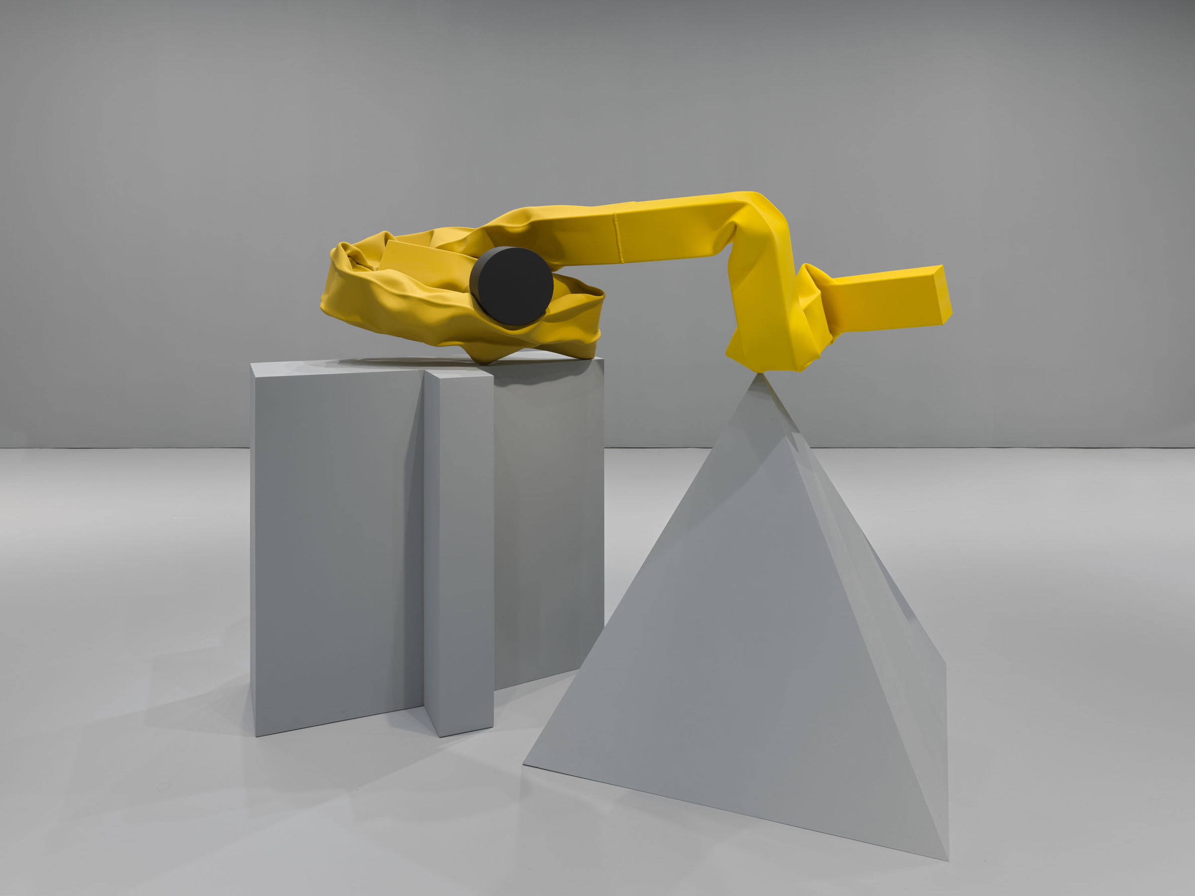 A yellow abstract sculpture rests on geometric grey blocks and a pyramid in a minimalist setting.