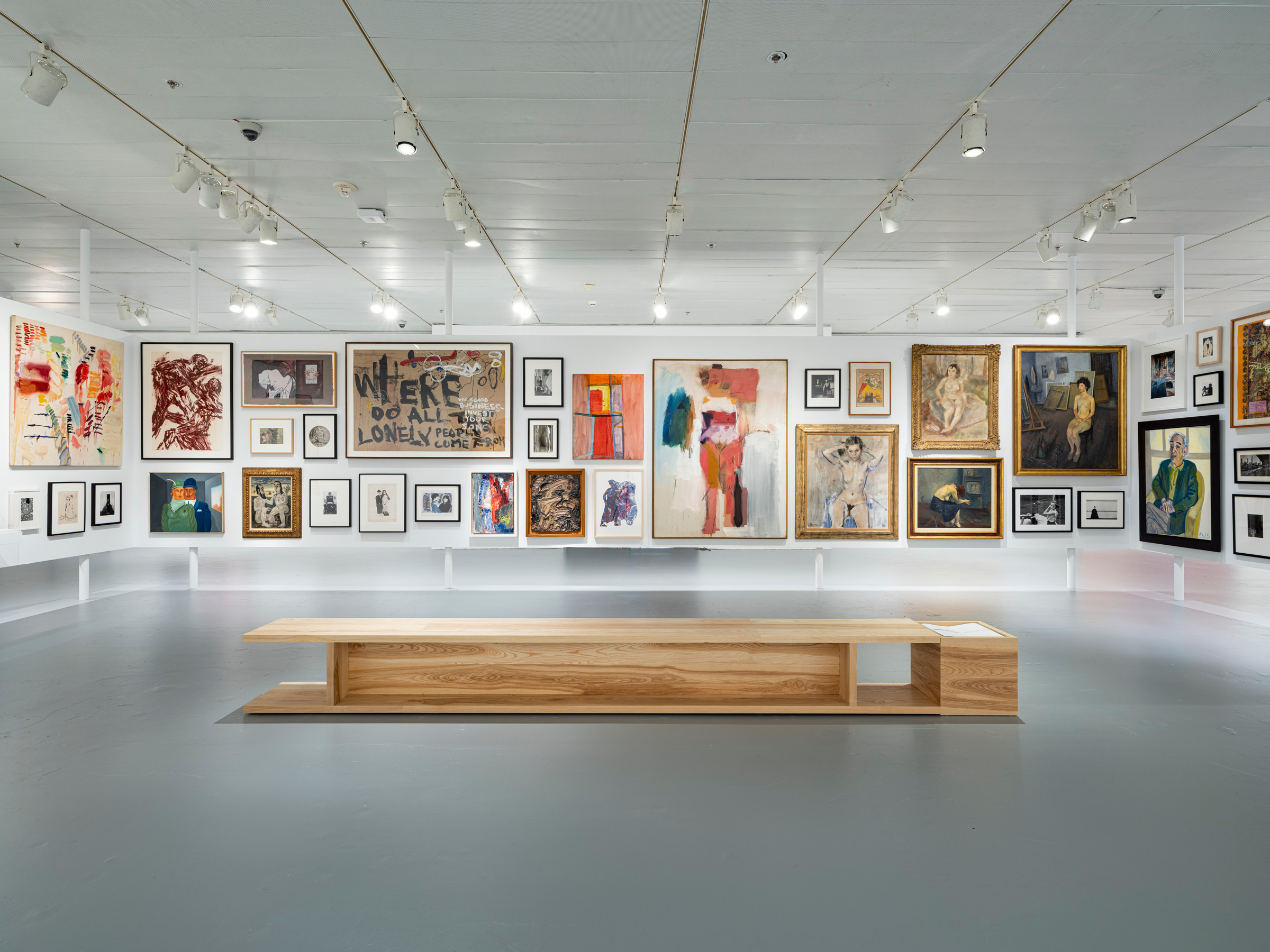A modern art gallery with numerous diverse paintings on white walls and a wooden bench in the center.