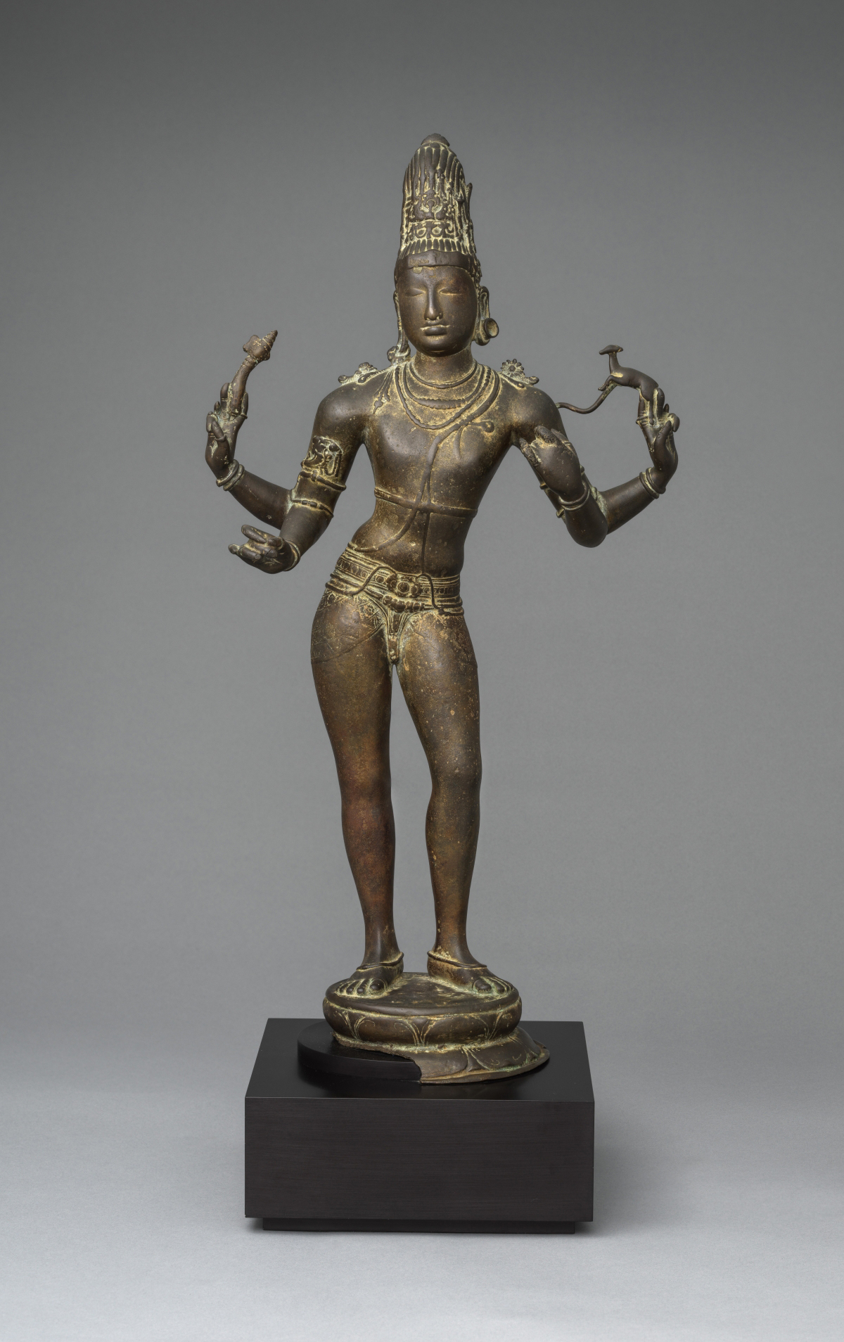 A bronze statue of a four-armed deity stands on a lotus pedestal, adorned with intricate jewelry.