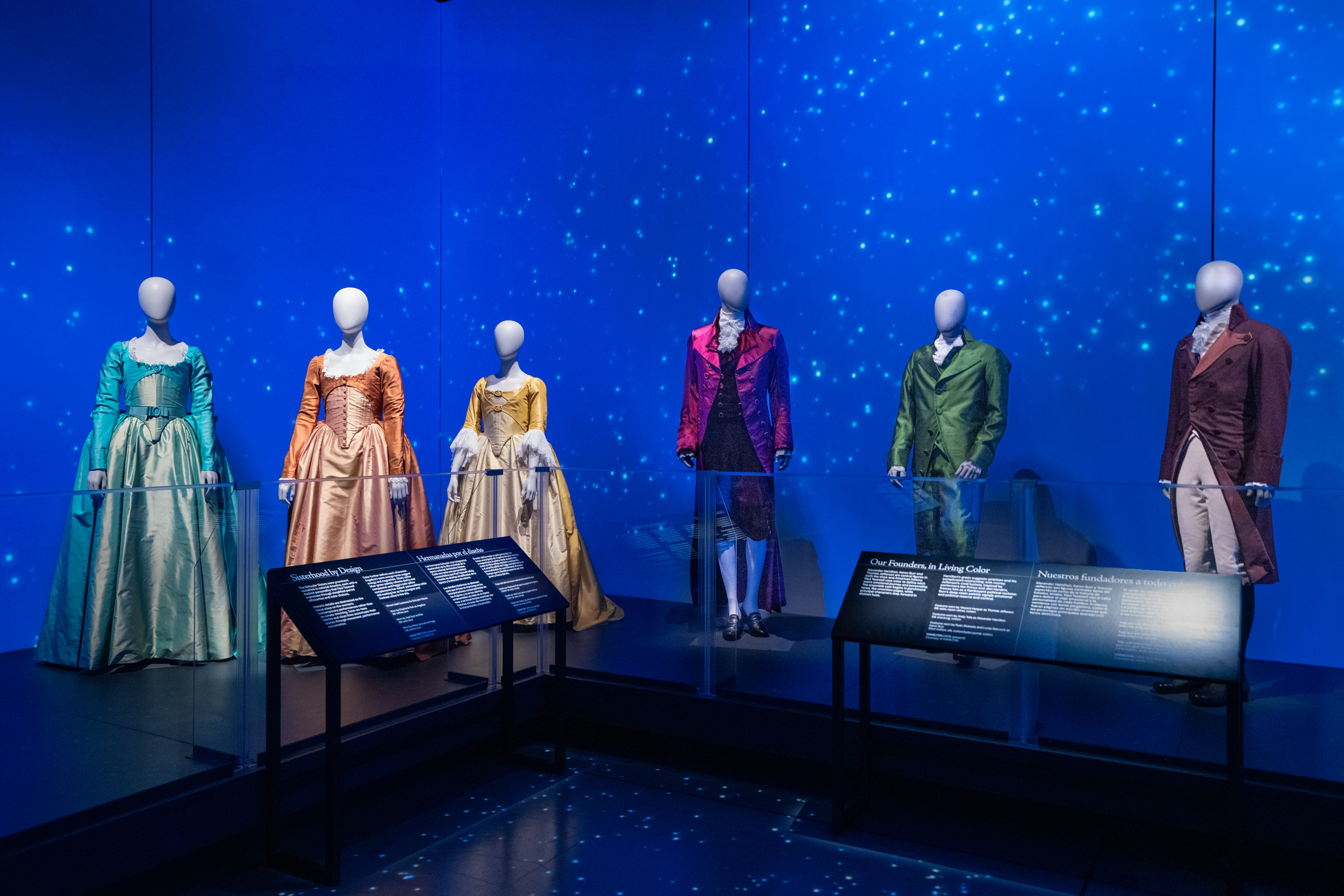 Mannequins in colorful period costumes are displayed against a blue, starry backdrop in a museum.