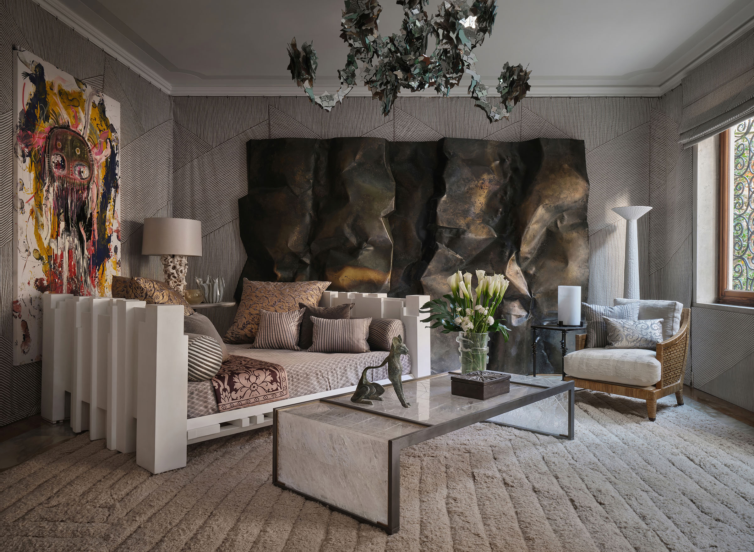 Among the artworks and design pieces in the boudoir at Fortuny + Chahan are, from left: Emilio Martinez's painting Life Totem, 2023; a unique lacquered wooden bed by Carla Venosta, 1972; a brass wall sculpture by Pierre Sabatier, 1998; a large Waterfall coffee table in rock crystal marquetry by Chahan; and a vintage McGuire rattan lounge chair. Overhead is a Saint-Just glass chandelier by Nathalie Ziegler. The walls are upholstered in Fortuny fabric