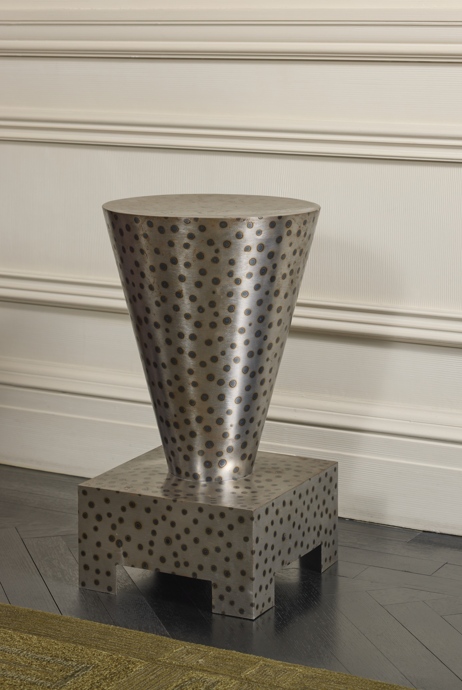 A metallic, cone-shaped stool with polka dot pattern sits on a square base against a wall.