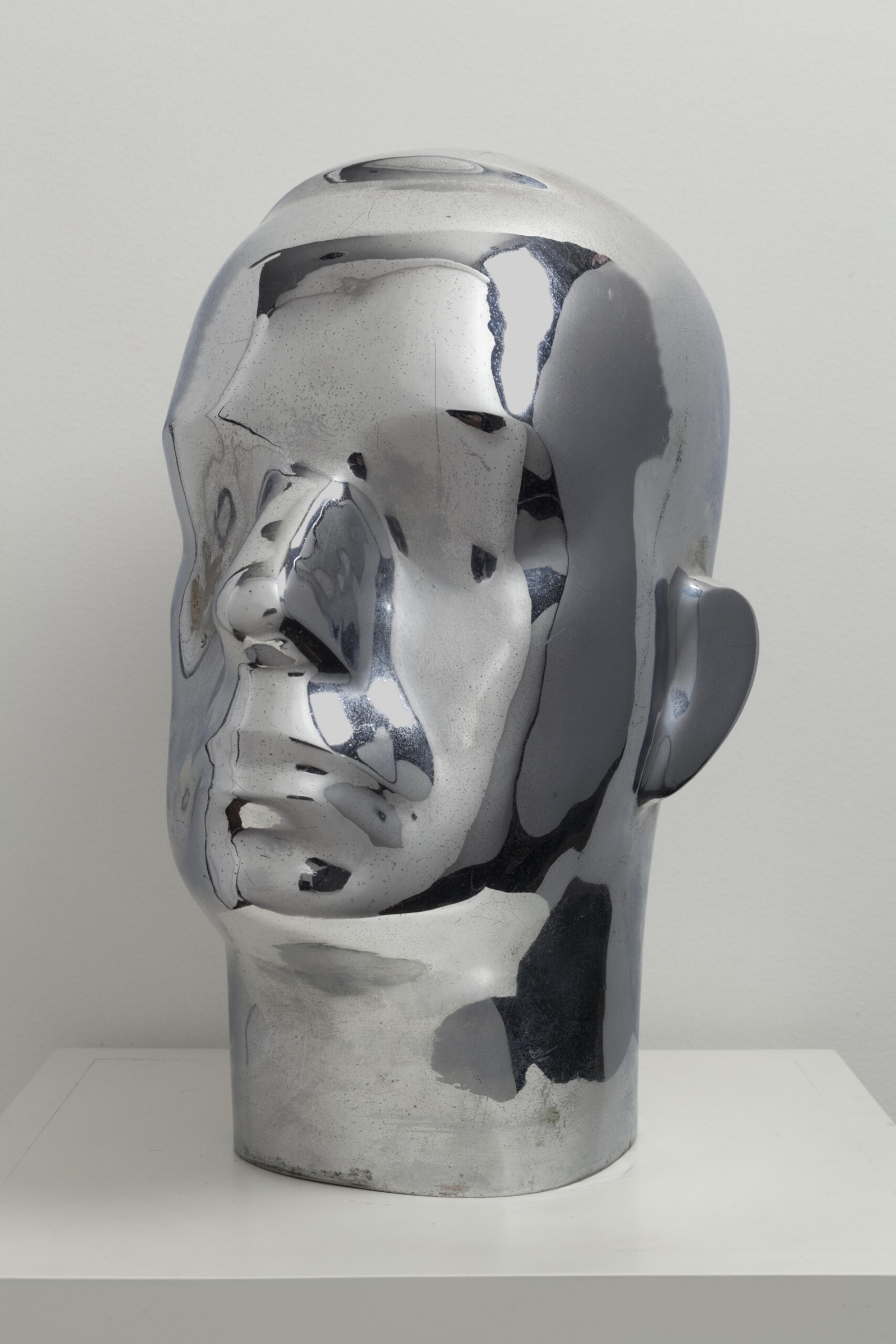 A shiny metallic head sculpture is displayed on a white pedestal against a plain background.