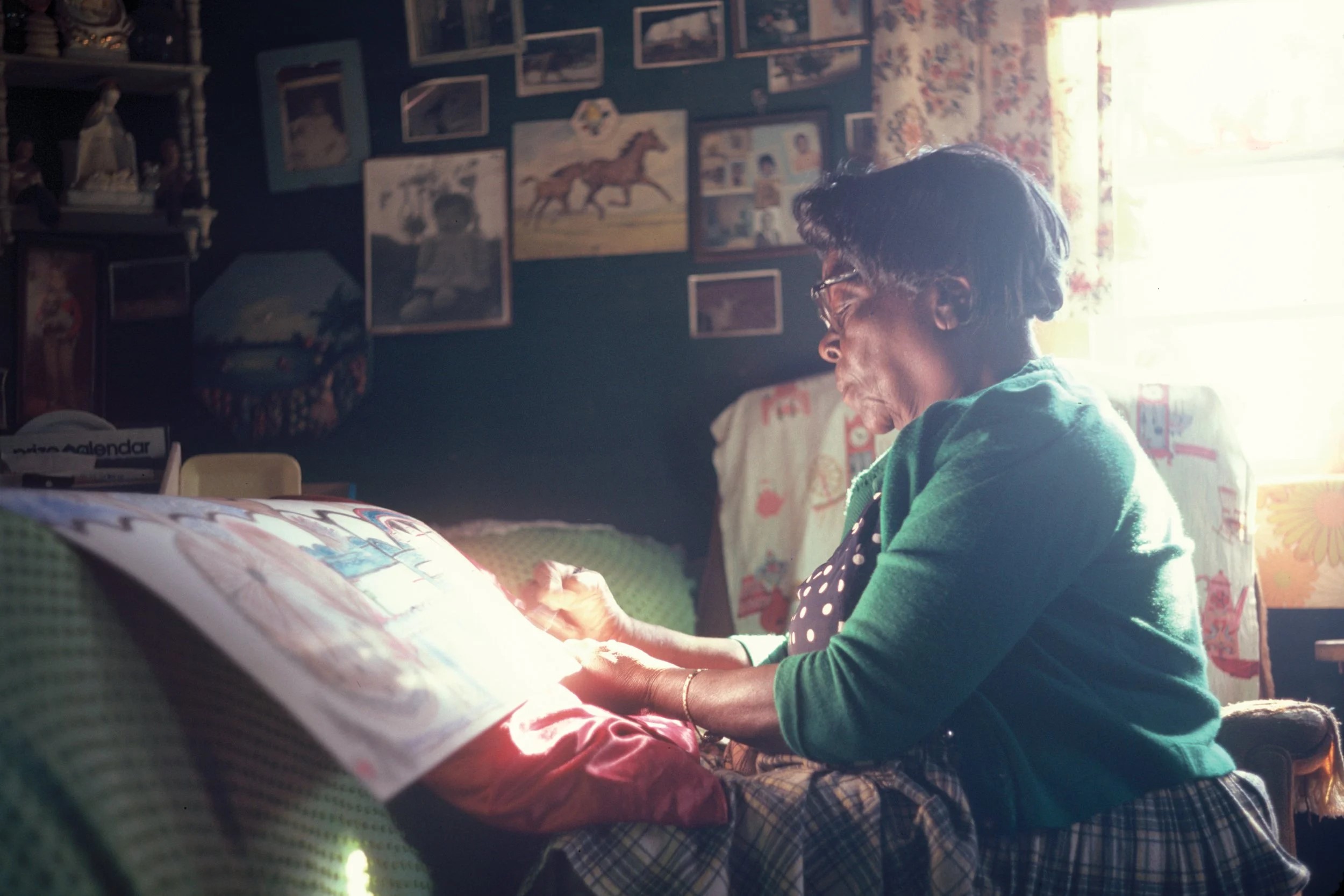A woman in a green sweater is drawing or painting while sitting in a cozy, sunlit room.