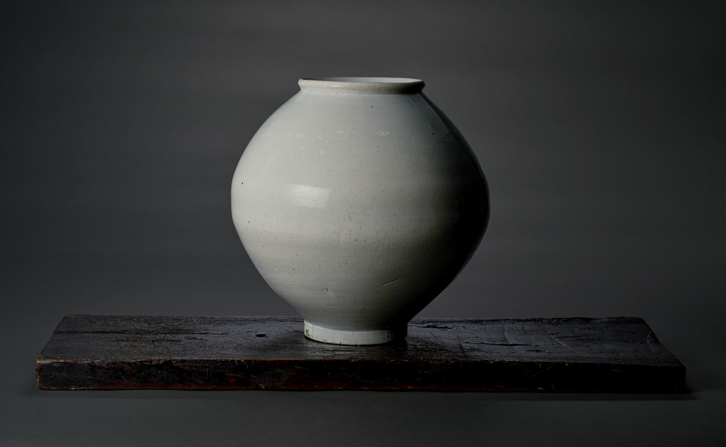 A round, white ceramic vase sits on a dark wooden surface against a gray background.