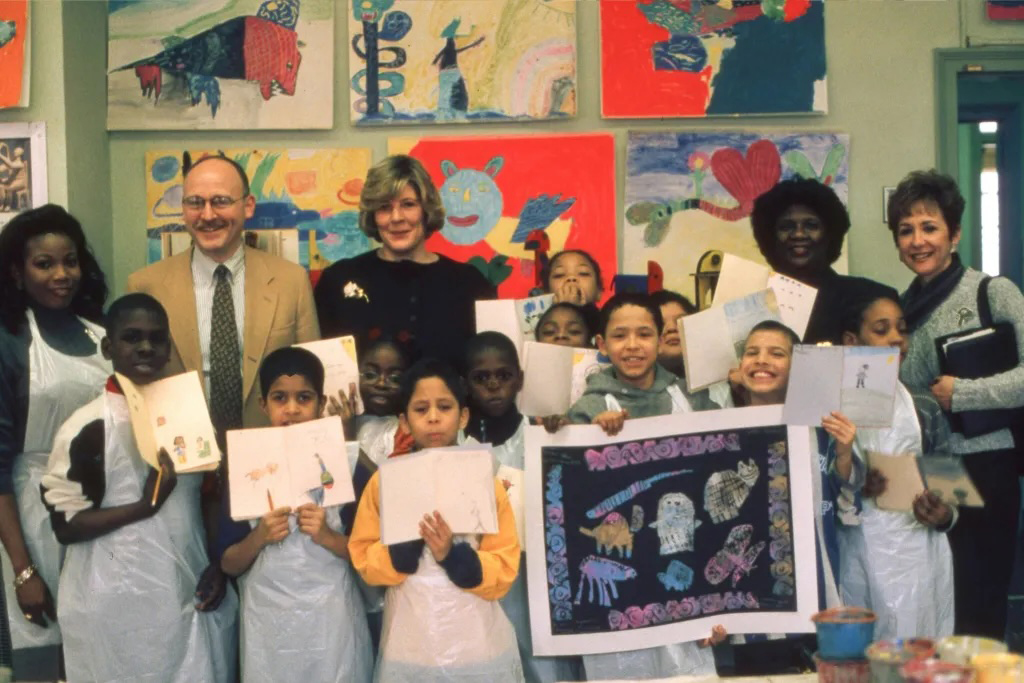Children and adults stand together holding artwork in a colorful classroom with paintings on the wall.