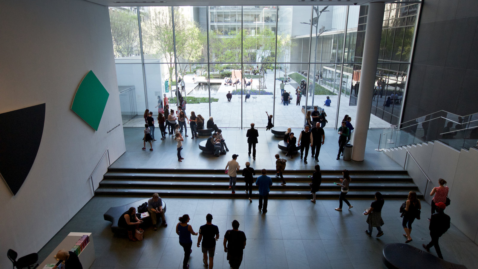 People walk and gather in a modern museum lobby with large windows overlooking a garden.