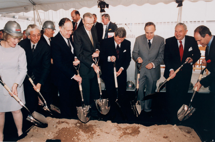 A group of people in formal attire are participating in a groundbreaking ceremony with shovels.