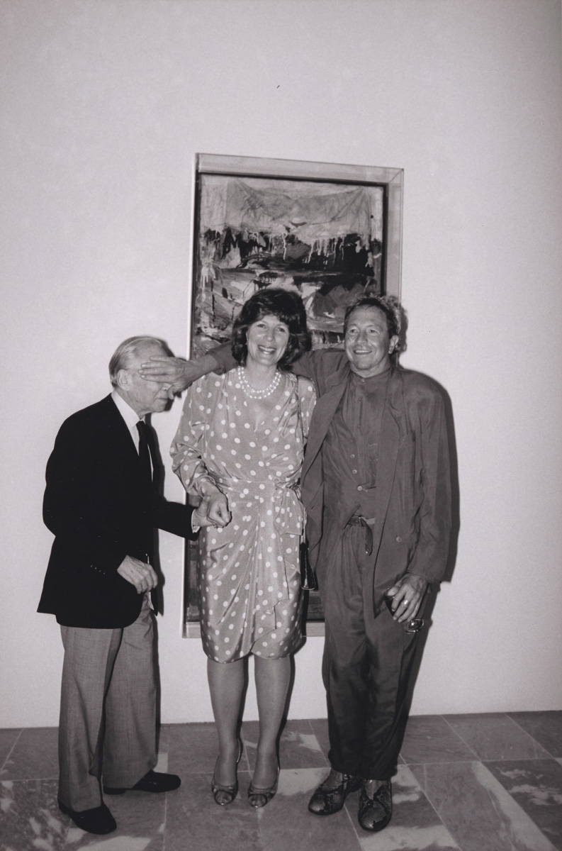 Three people stand together in front of a framed painting, posing for a photo in formal attire.