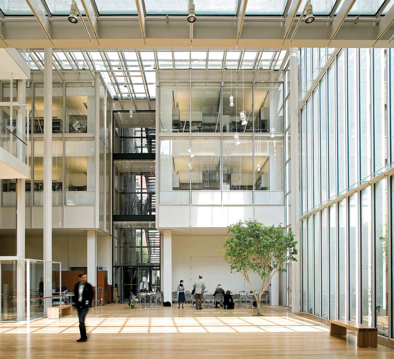 A spacious modern atrium with glass walls, wooden floors, trees, and people working inside.