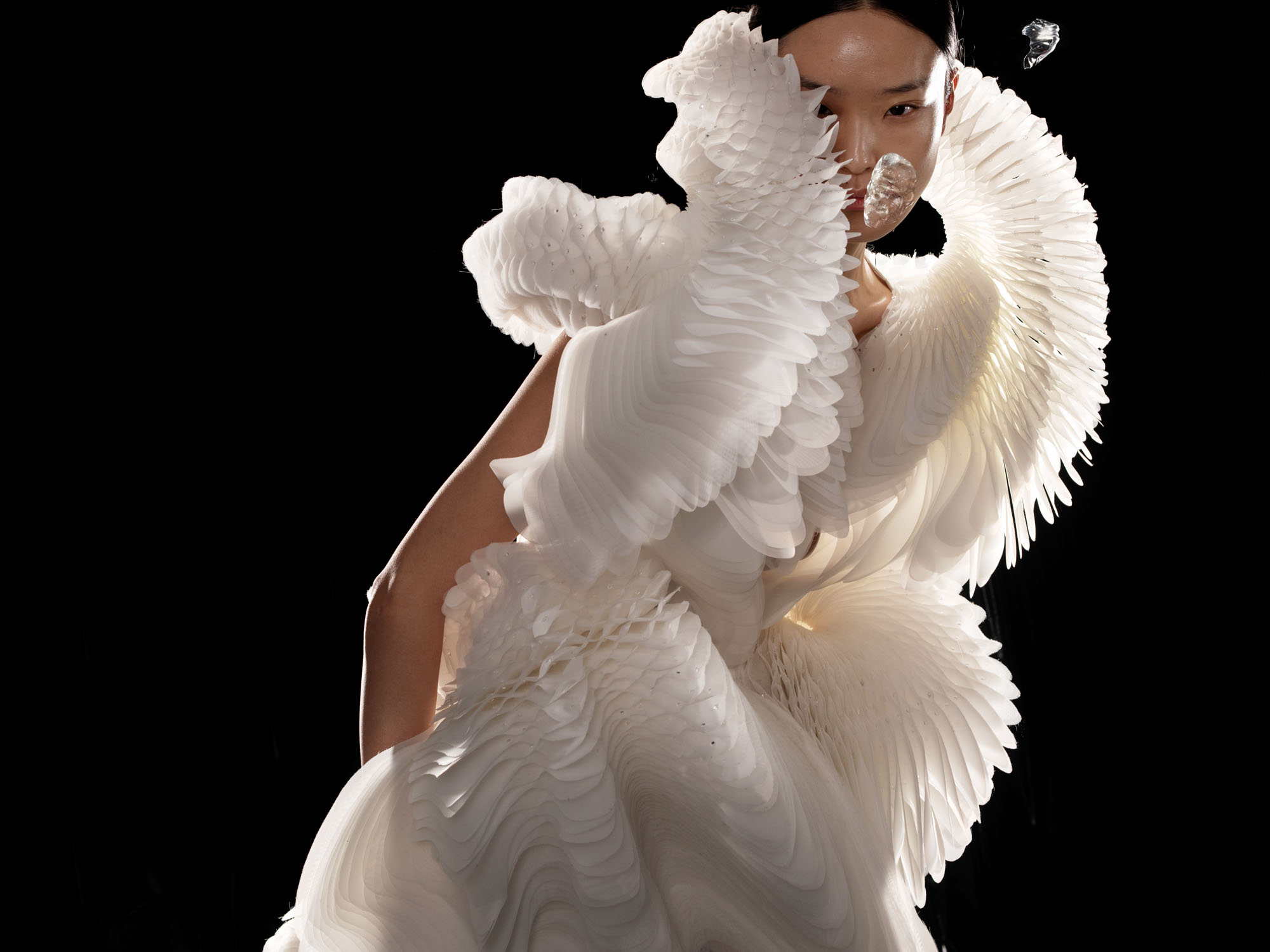 A person wearing an elaborate white sculptural dress with feather-like textures stands against a black background.