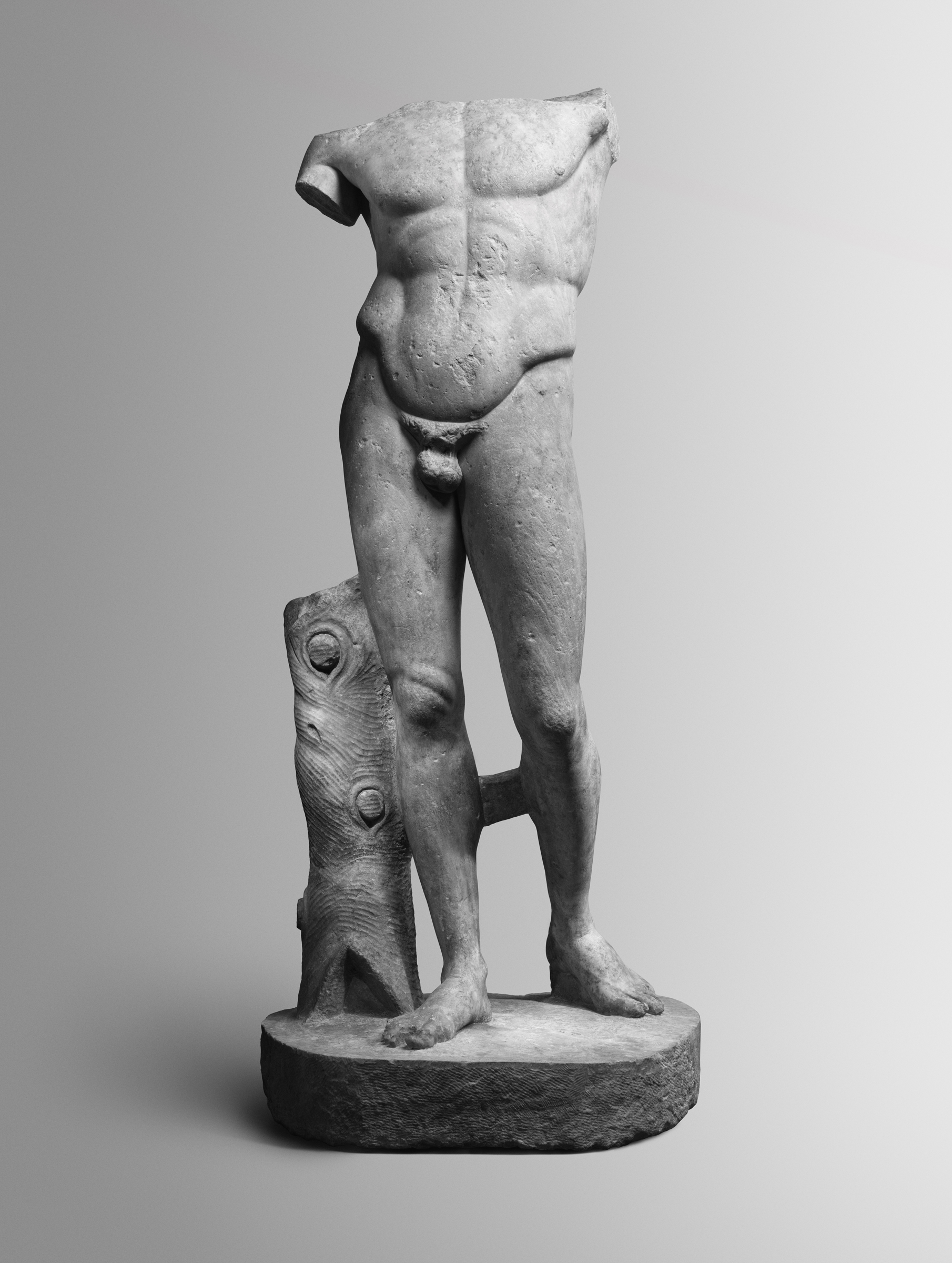 A headless and armless ancient marble statue of a male figure stands on a round base.