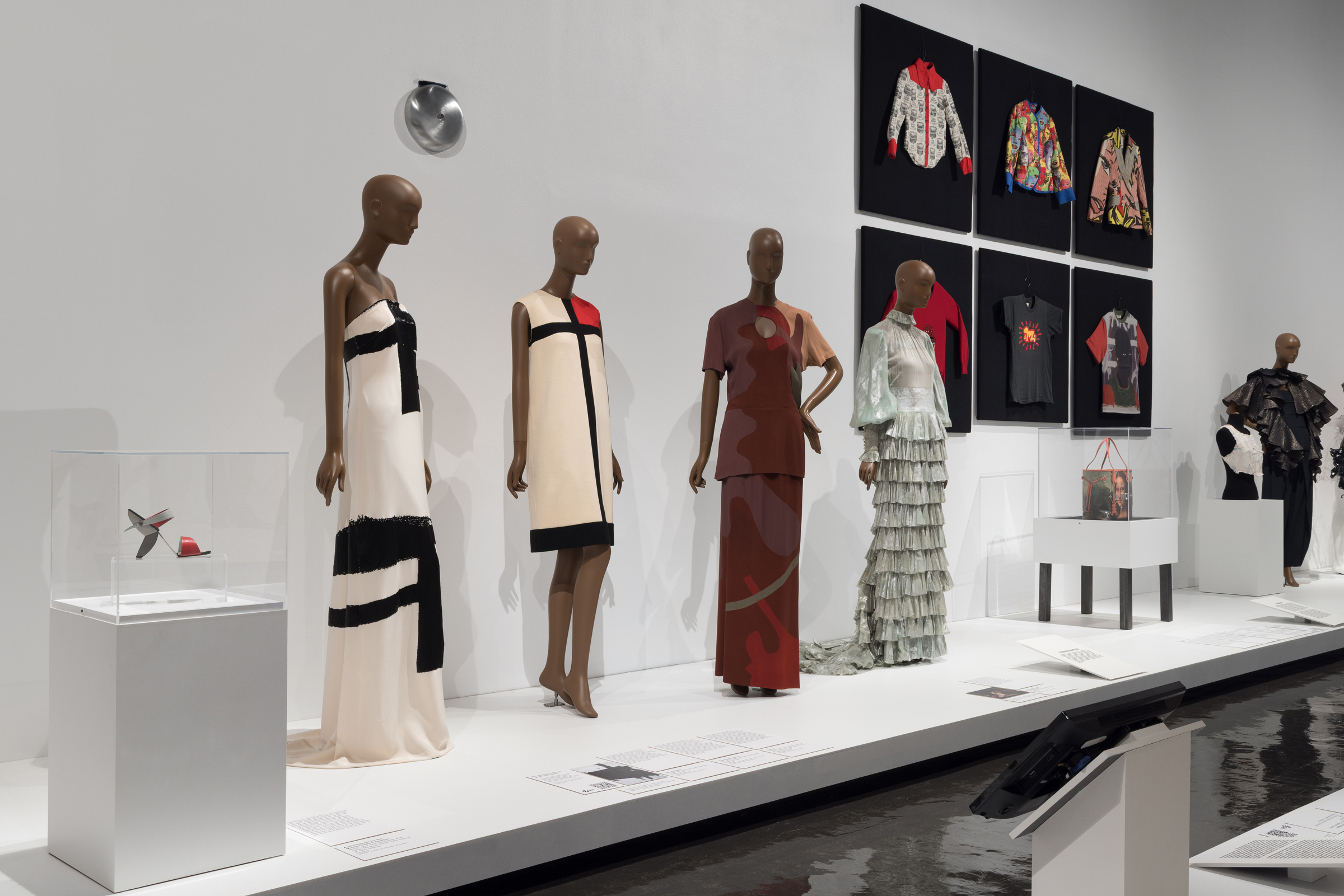Four mannequins display stylish dresses with framed garments and accessories on the wall behind them.