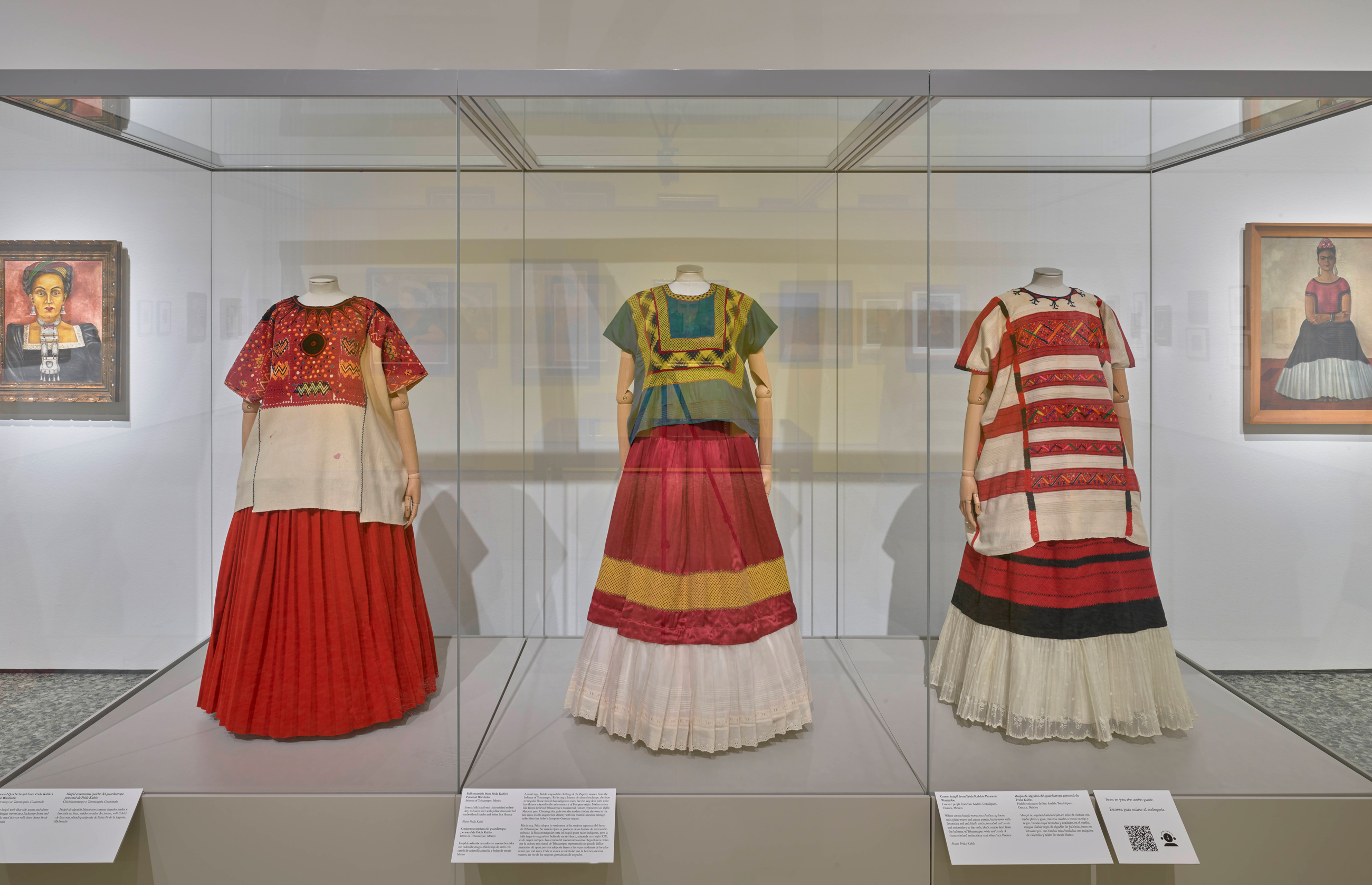 Three colorful traditional dresses are displayed in glass cases at a museum exhibit.