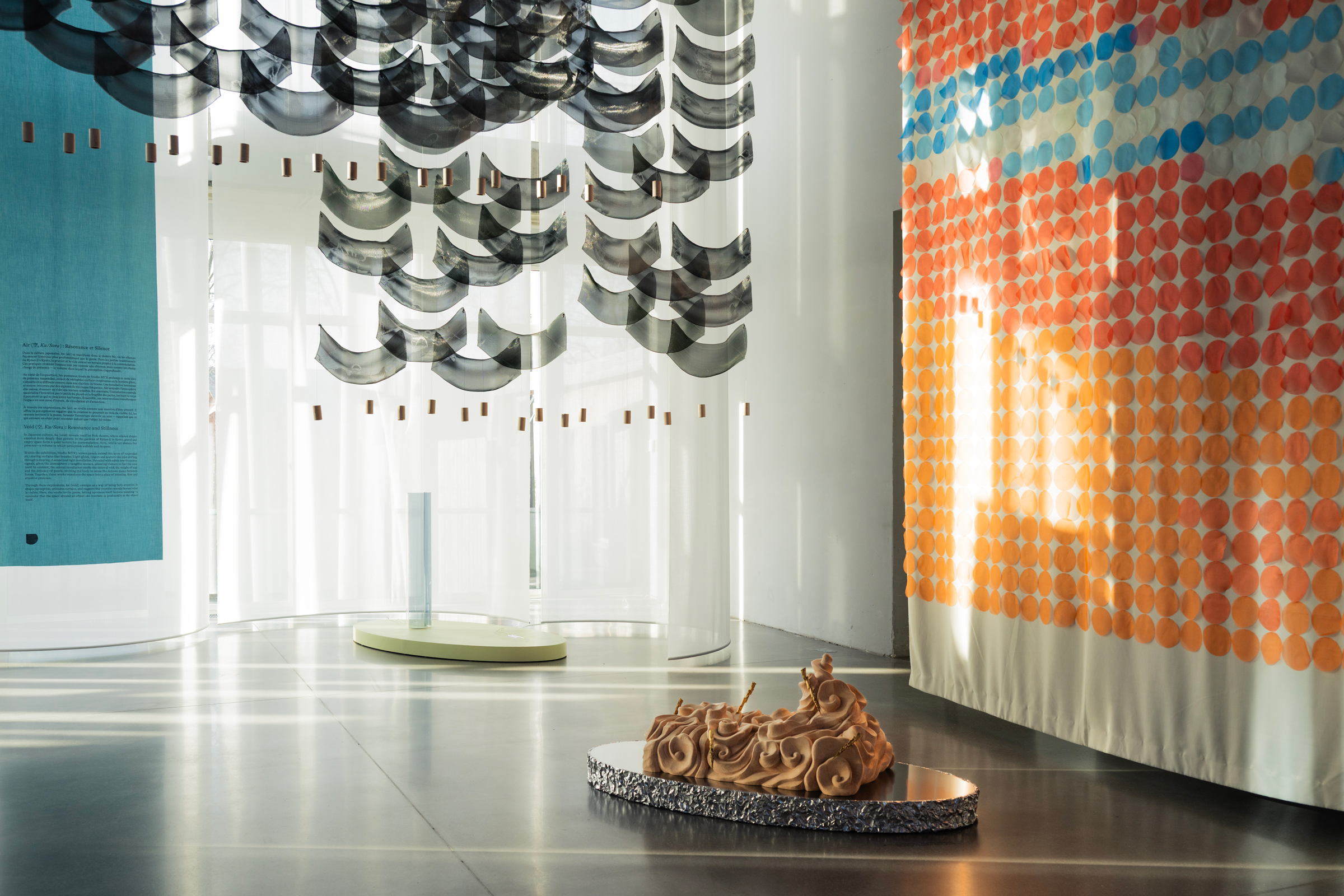 Modern art installation with hanging fabric, colorful dot panels, and a sculpted object on the floor.