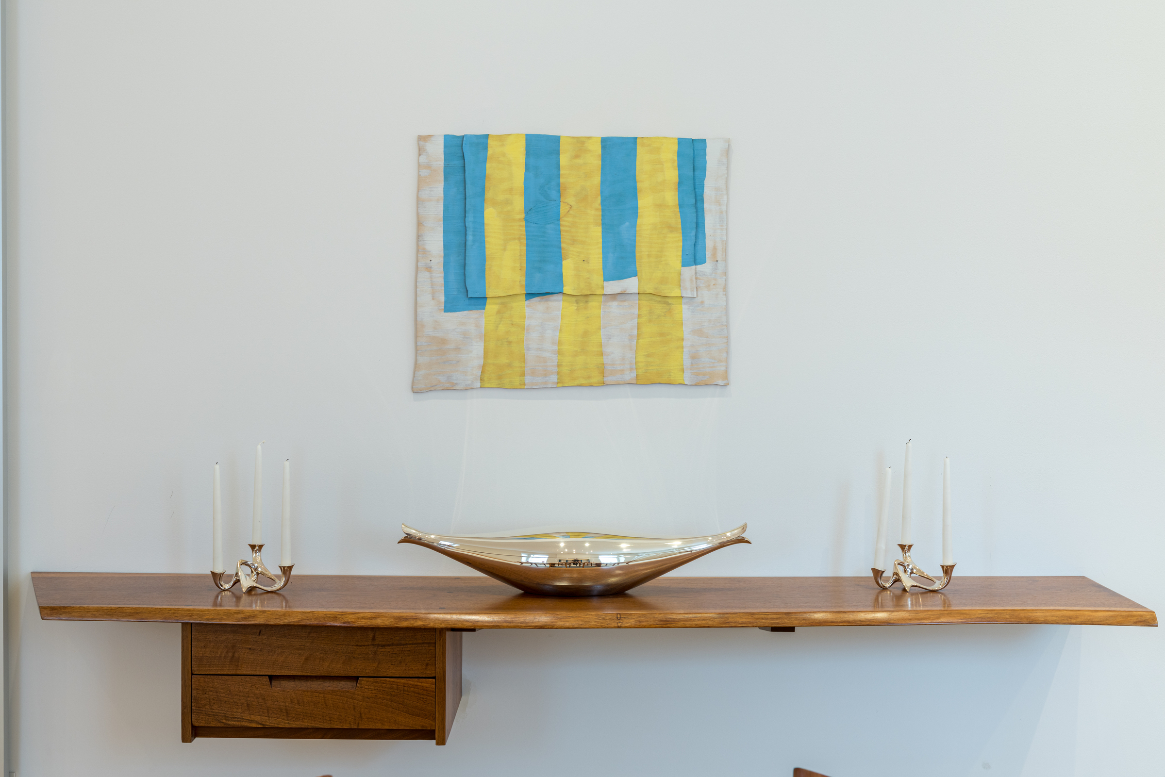 A wooden shelf with candle holders, a decorative bowl, and abstract art with blue and yellow stripes.