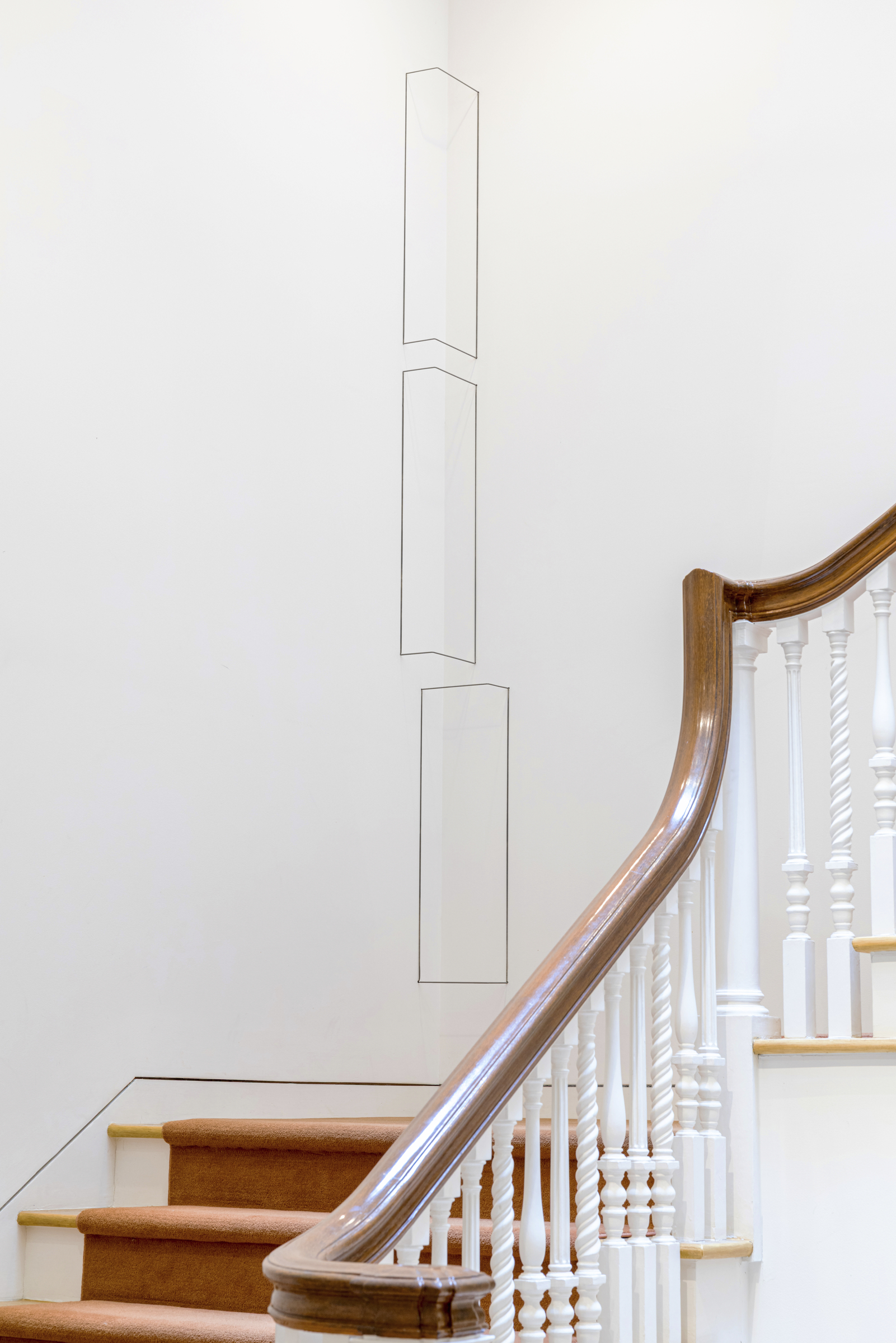 Three outlined geometric shapes are drawn on a white wall above a staircase with a brown handrail.