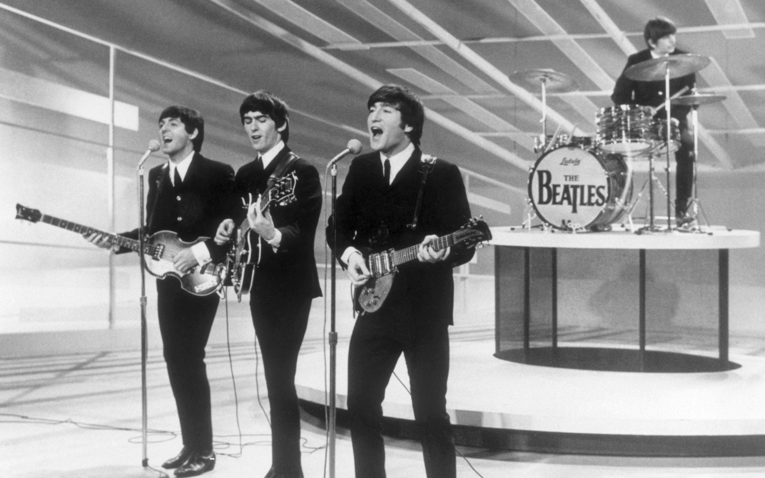 Four musicians in suits perform on stage with instruments and a drum set labeled "The Beatles."