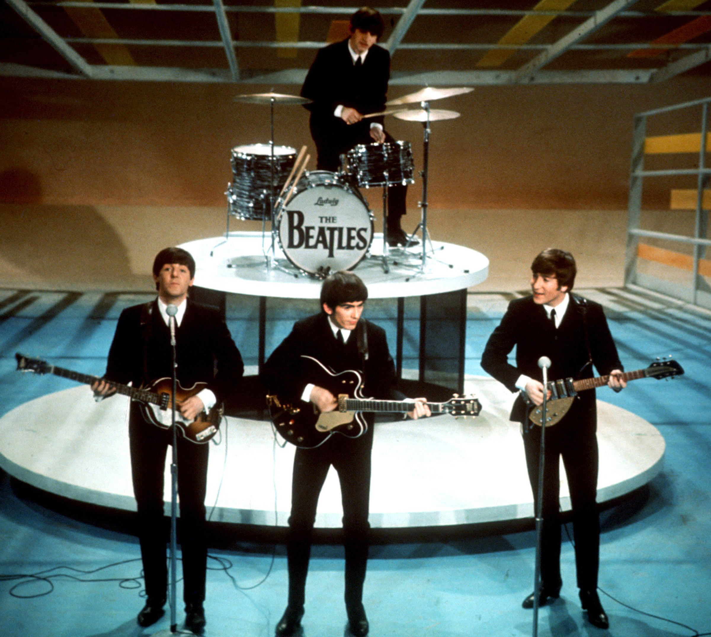 Four musicians in suits perform on stage with guitars and drums labeled "The Beatles."