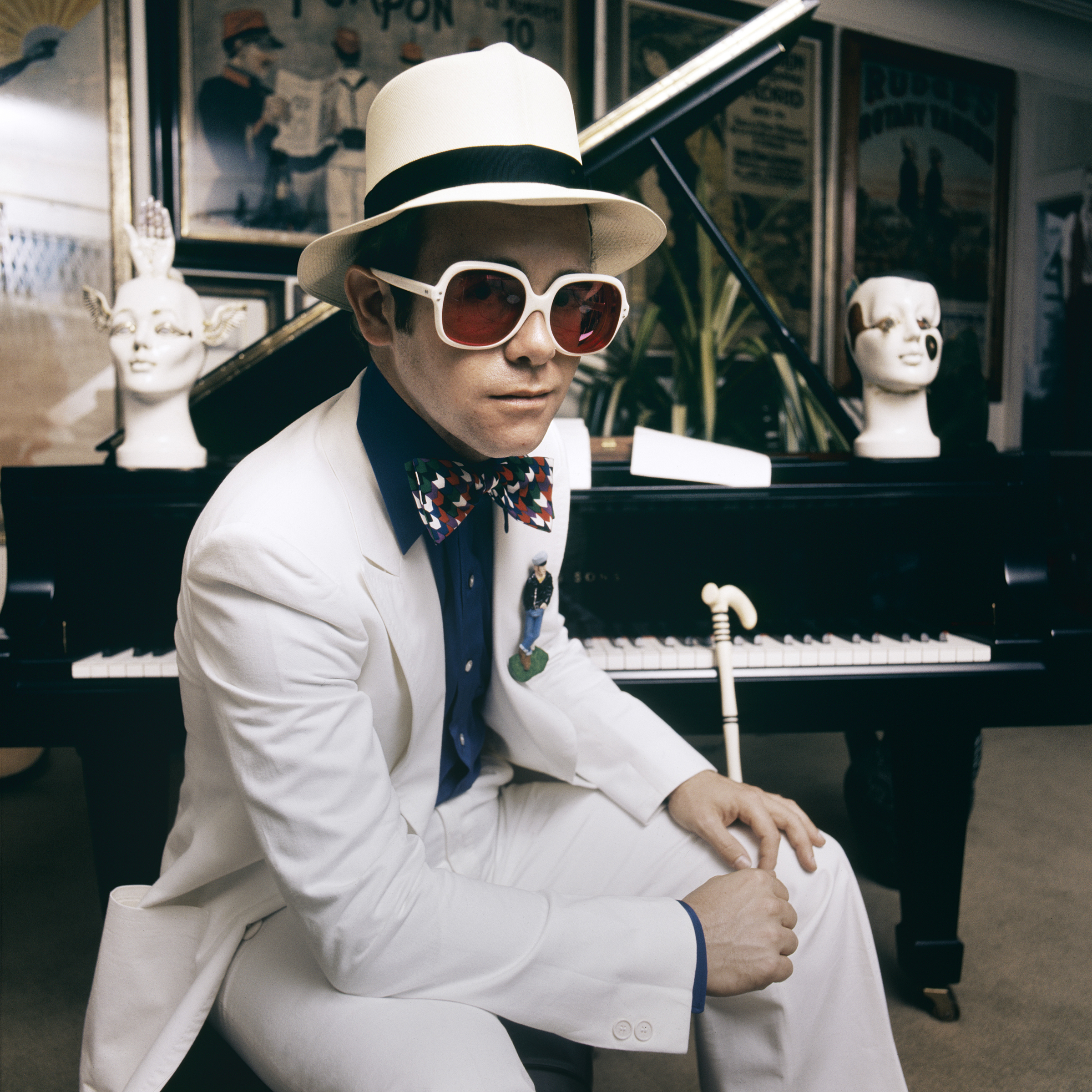 A person in a white suit and hat sits beside a grand piano with posters and plants in the background.
