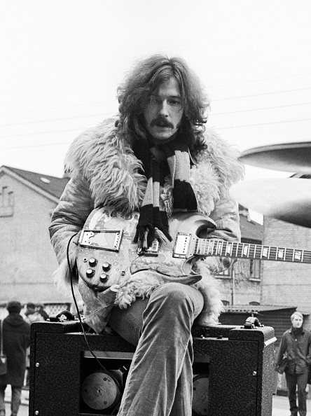 A person wearing a fur coat and scarf is sitting on an amplifier with an electric guitar outdoors.