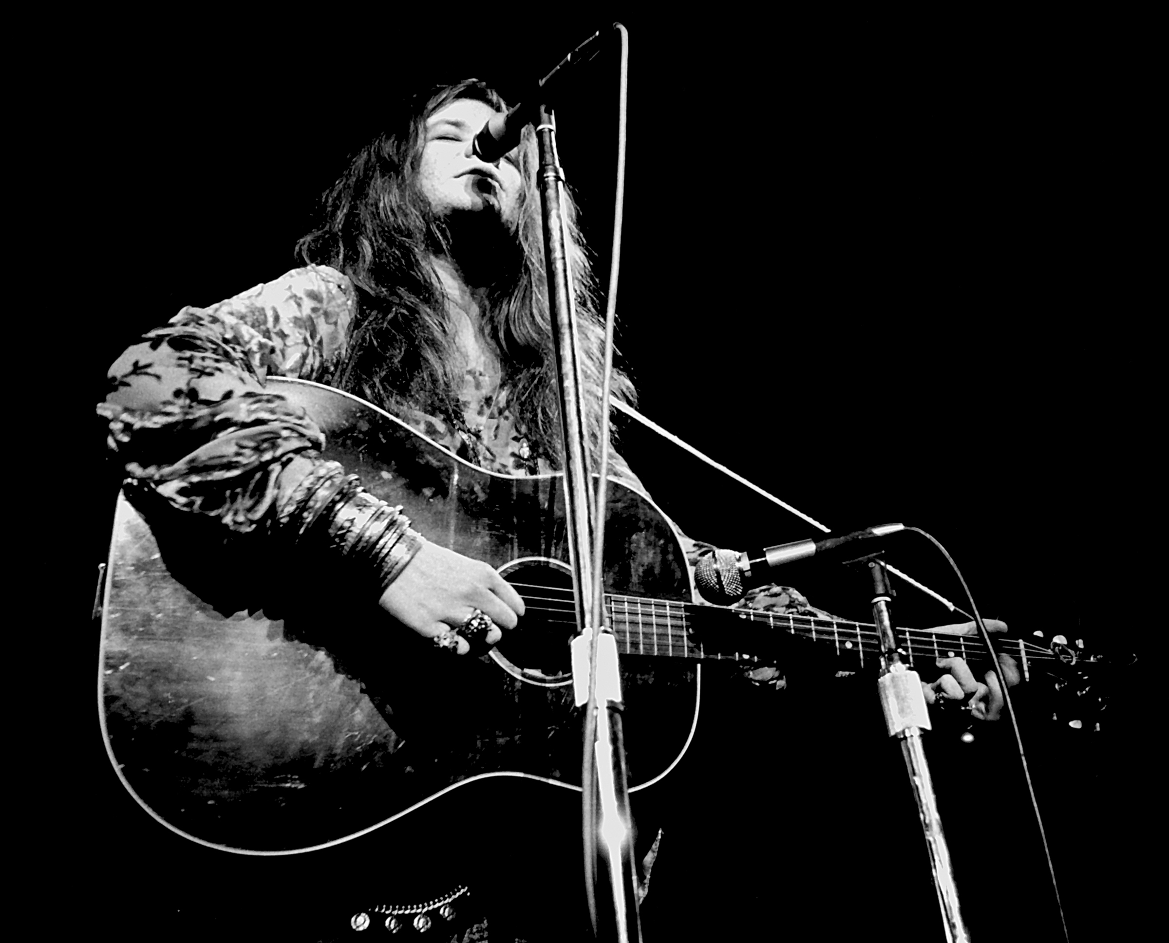 A person with long hair and bracelets plays an acoustic guitar and sings into a microphone.