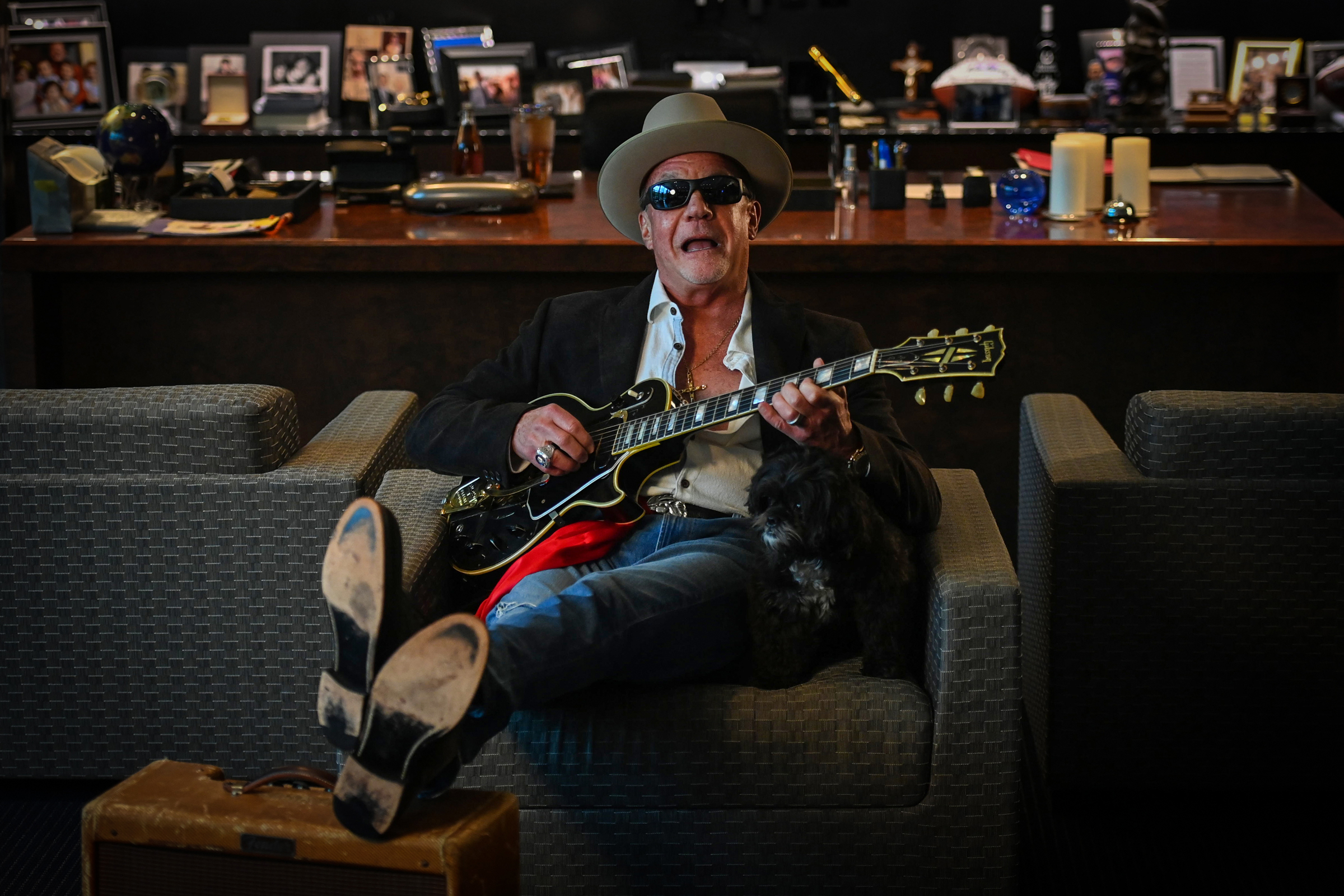 A person wearing a hat plays guitar while sitting on a chair with a dog beside them.