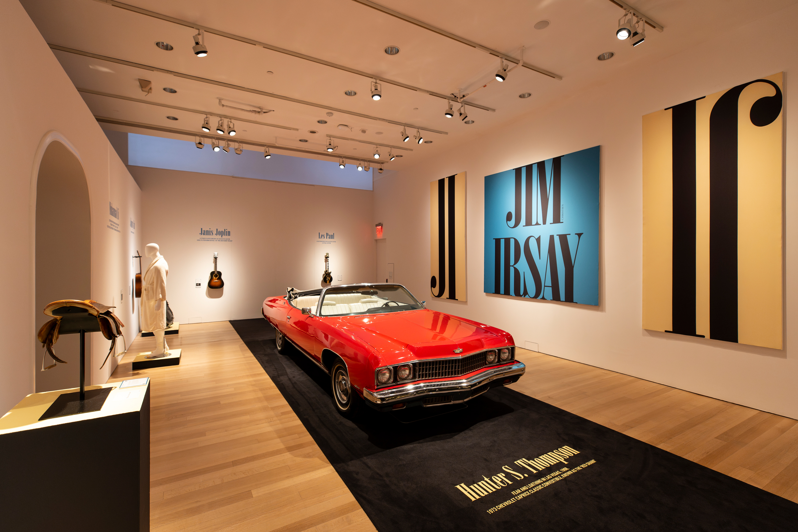 A red vintage convertible car is displayed in a modern gallery with guitars and art on the walls.