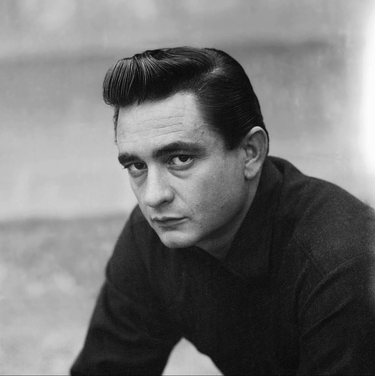 A person with slicked-back dark hair is wearing a black shirt in a black and white photo.